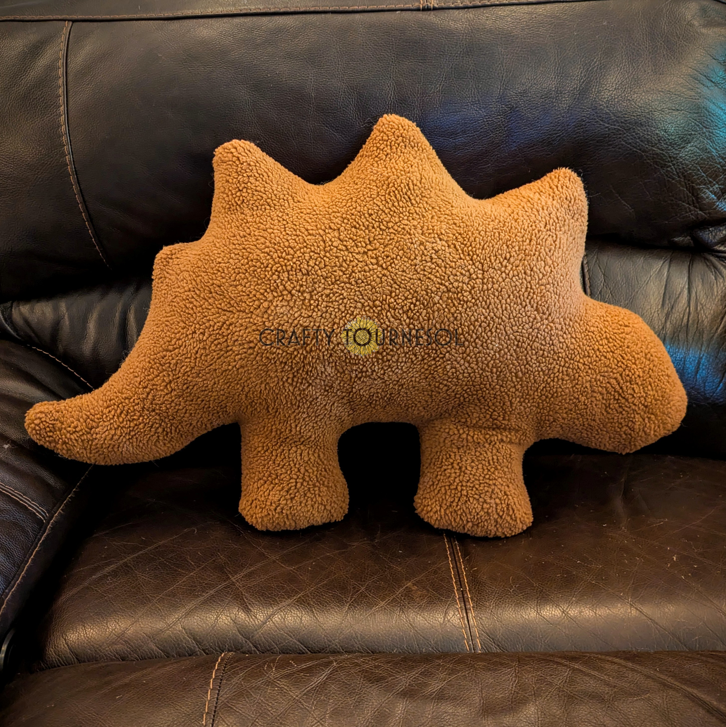 A 3-feet-long, medium brown sherpa pillow in the shape of a stegosaurus chicken nugget. Posed on a black leather sofa.