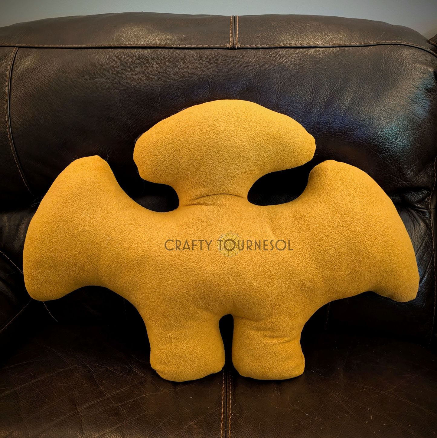A 3-feet-long, golden brown fleece pillow in the shape of a pterodactyl chicken nugget. Posed on a black leather sofa.