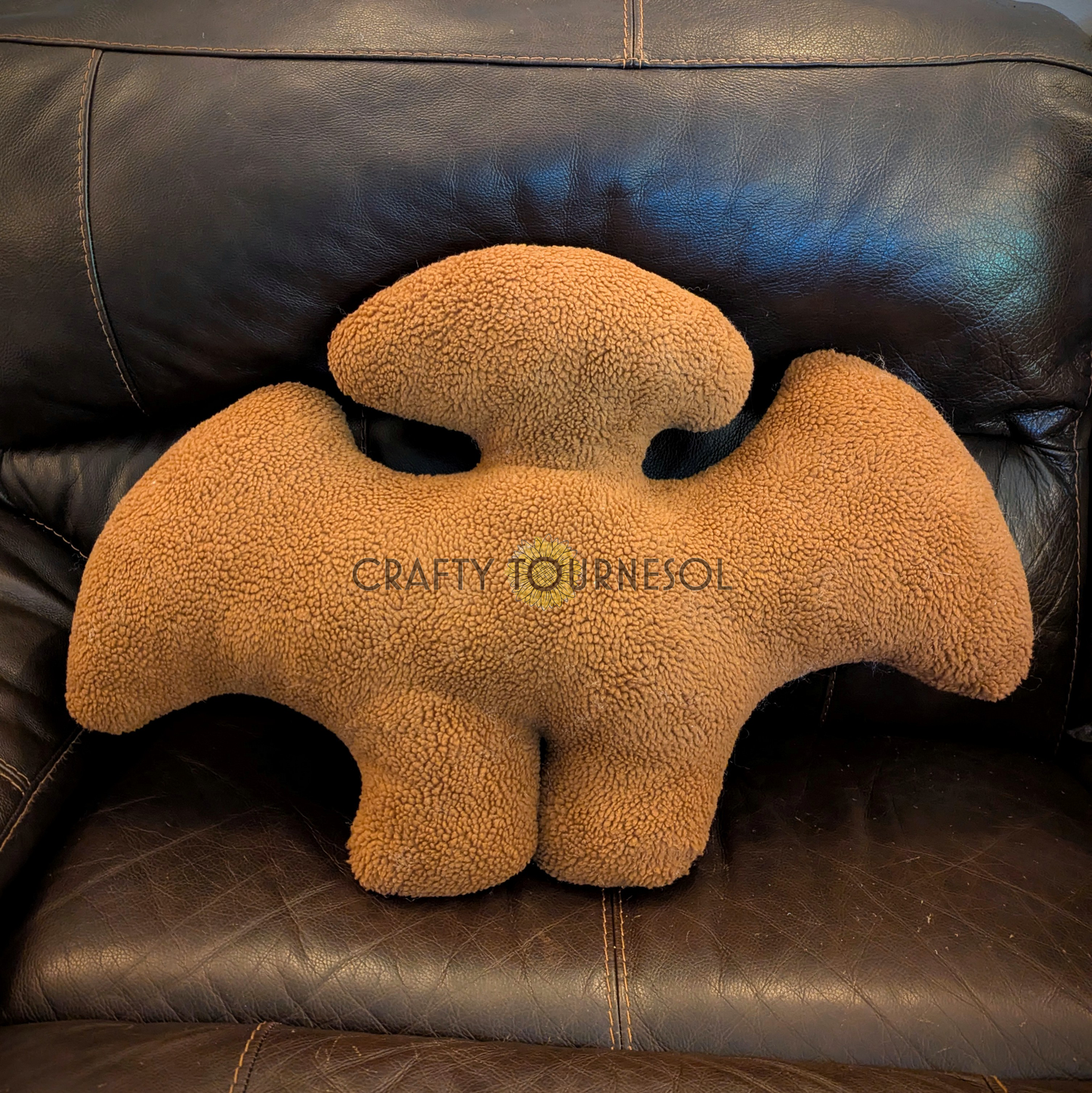 A 3-feet-long, medium brown sherpa pillow in the shape of a pterodactyl chicken nugget. Posed on a black leather sofa.