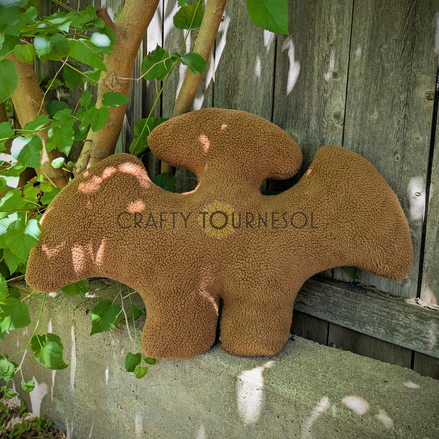 A 3-feet-long, medium brown sherpa pillow in the shape of a pterodactyl chicken nugget. Posed in front of a wooden fence and tree.