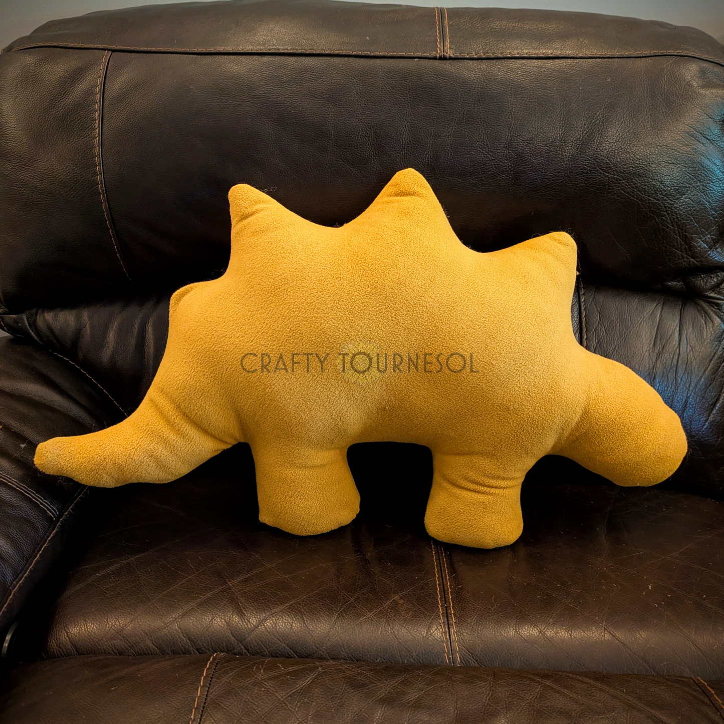 A 3-feet-long, golden brown fleece pillow in the shape of a stegosaurus chicken nugget. Posed on a black leather sofa.