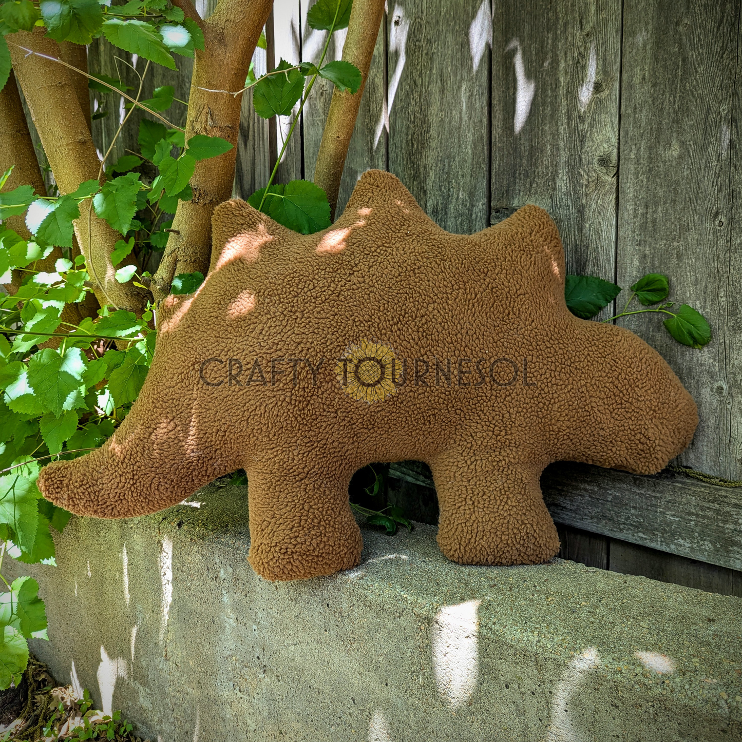 A 3-feet-long, medium brown sherpa pillow in the shape of a stegosaurus chicken nugget. Posed in front of a wooden fence and tree.
