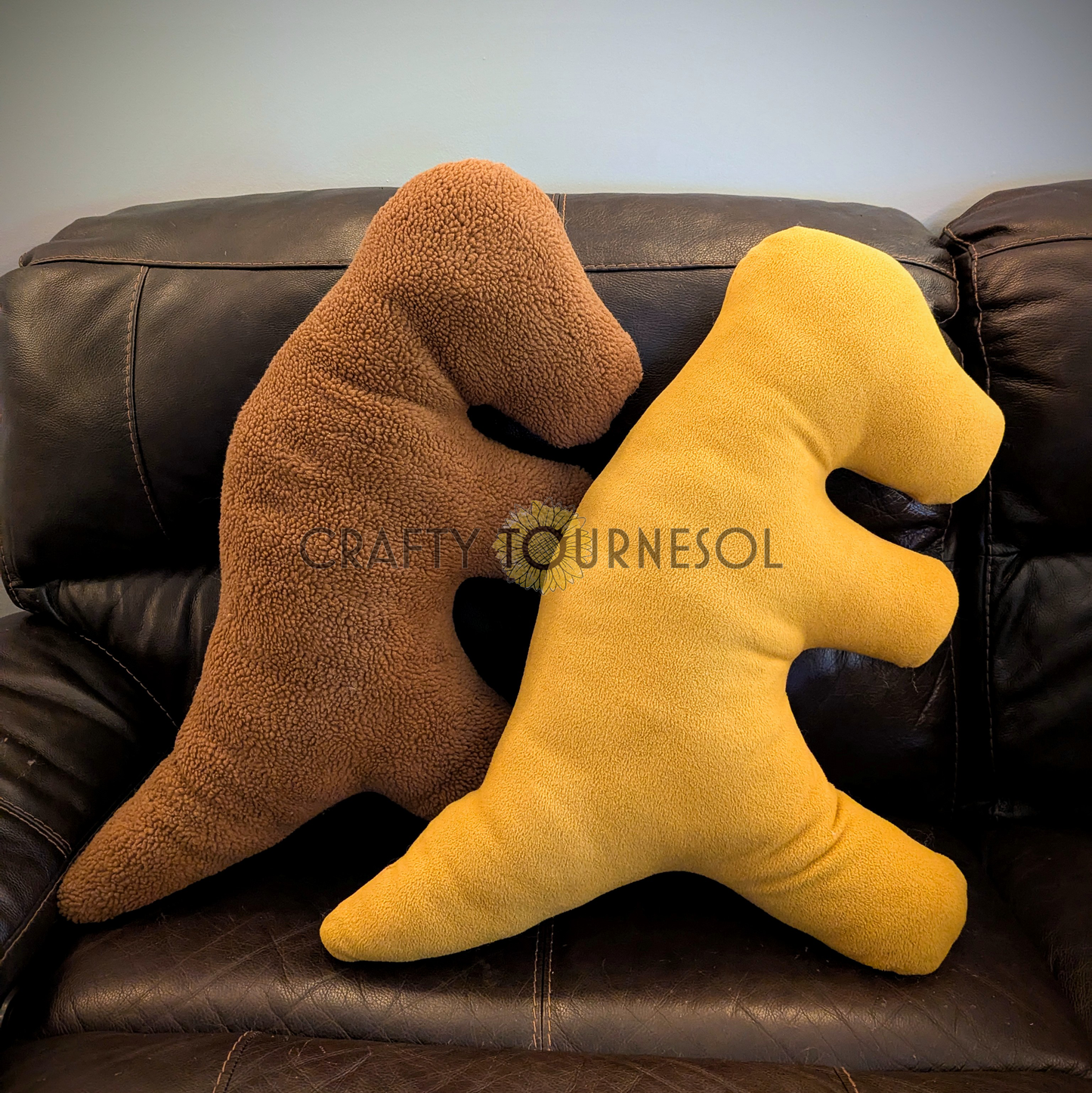 Two 3-feet-long pillows in the shape of t-rex dinosaur chicken nuggets. One is made with medium brown sherpa fabric, and the other is made with golden brown fleece fabric. Posed on a black leather sofa.