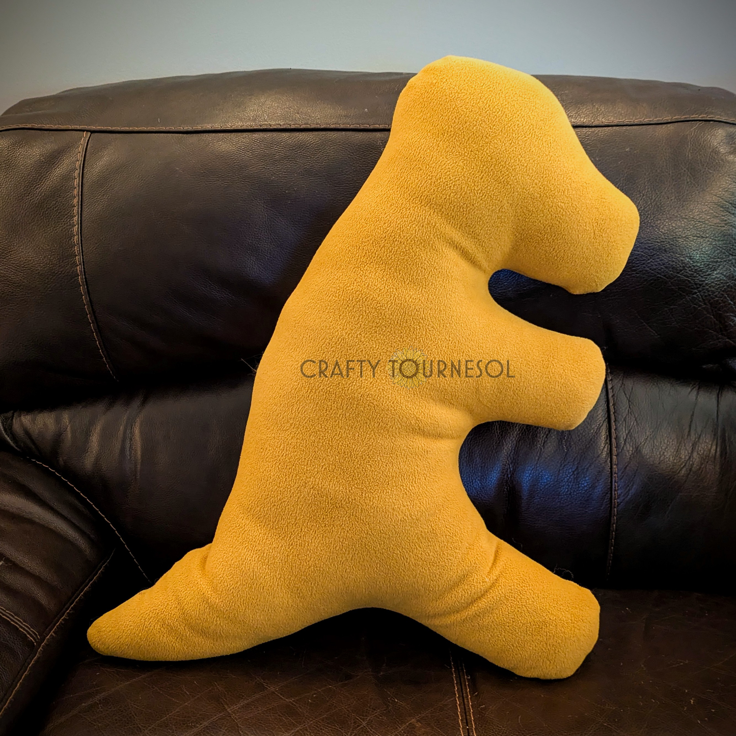 A 3-feet-long, golden brown fleece pillow in the shape of a t-rex chicken nugget. Posed on a black leather sofa.