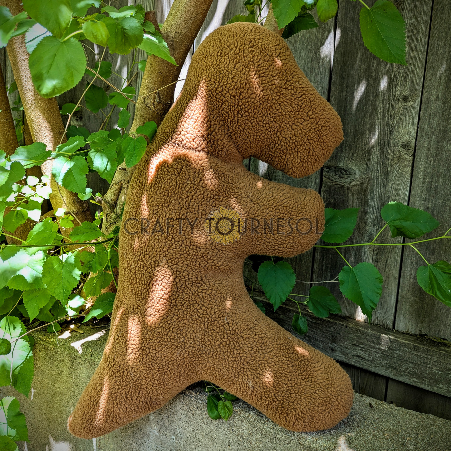 A 3-feet-long, medium brown sherpa pillow in the shape of a t-rex chicken nugget. Posed in front of a wooden fence and tree.