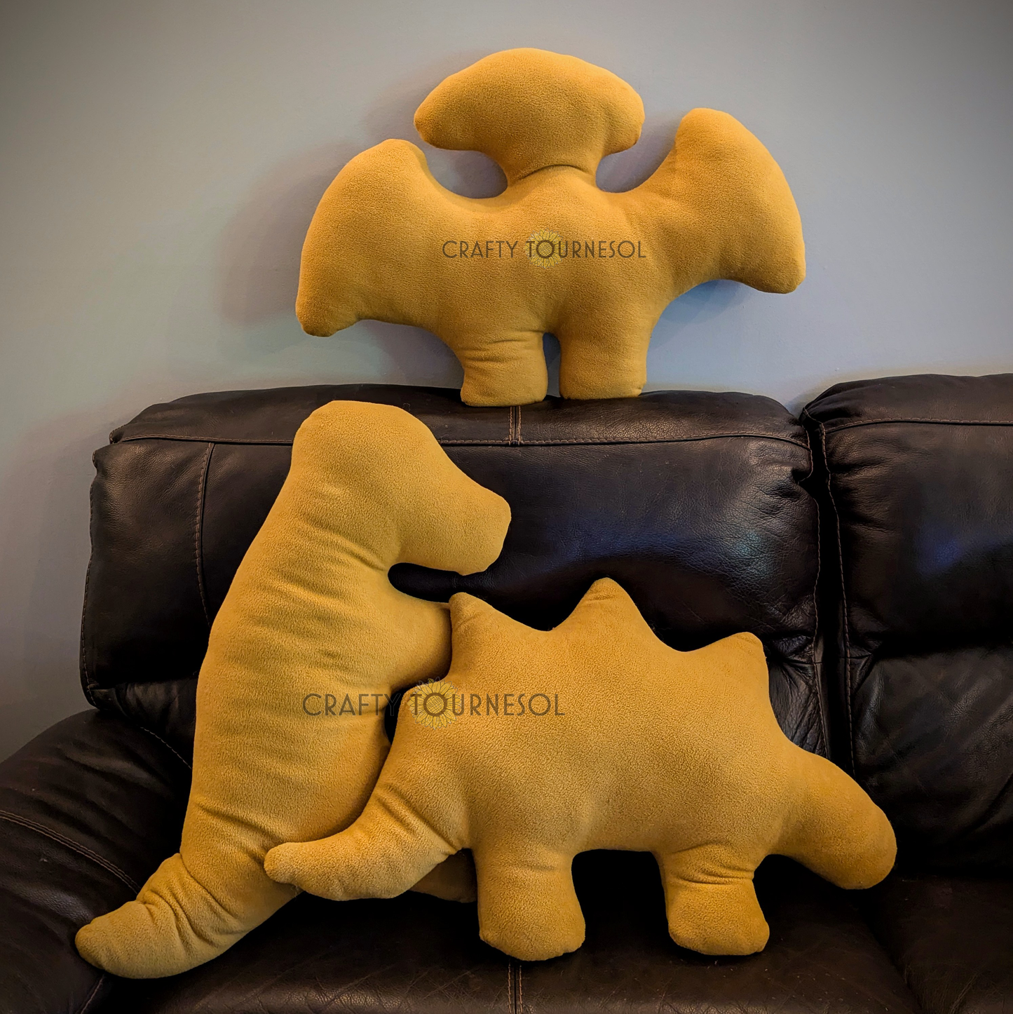 Three 3-feet-long, golden brown fleece pillows in the shape of dinosaur chicken nuggets. A t-rex, stegosaurus, and pterodactyl. Posed on a black leather sofa.