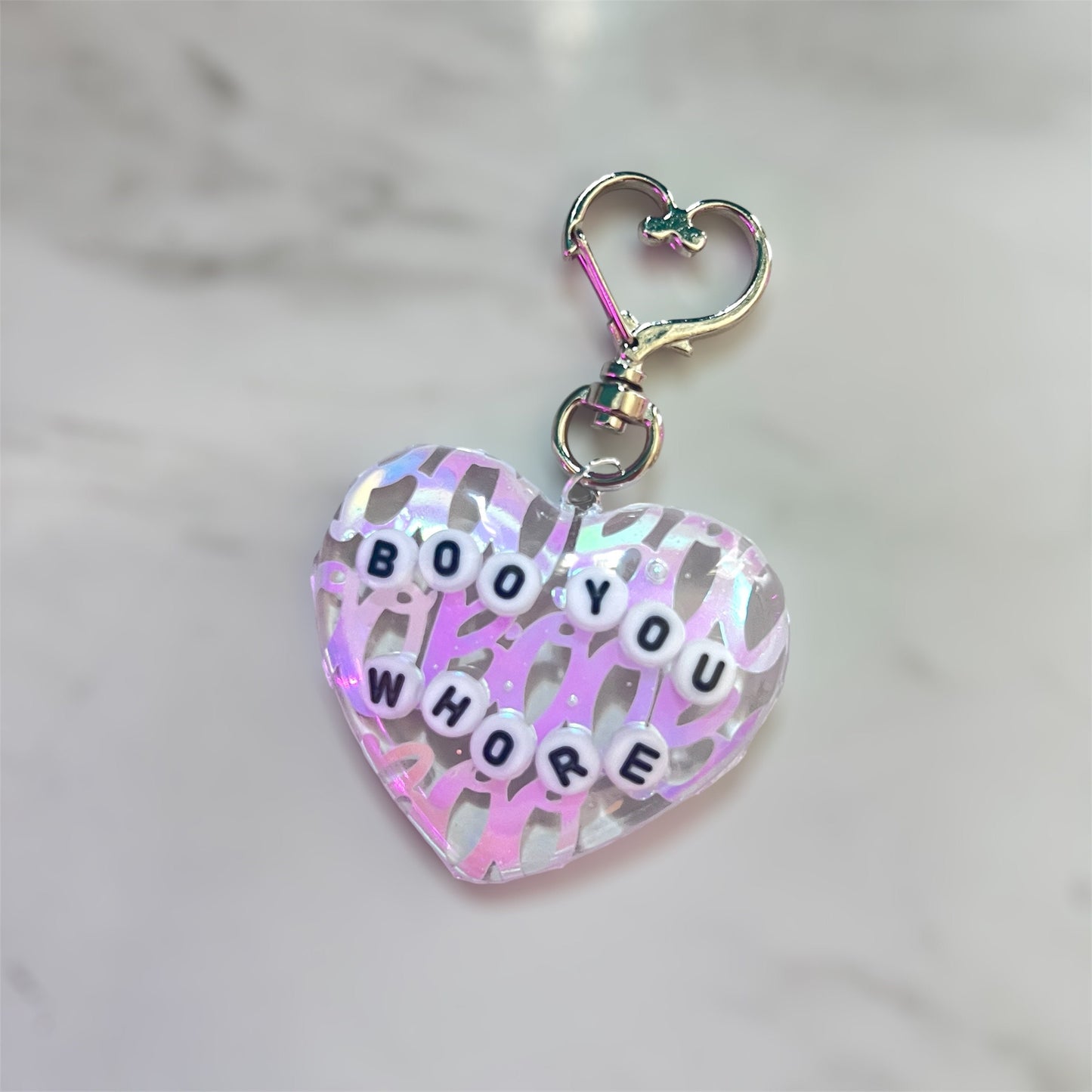 Heart-shaped resin keychain with the phrase 'Boo You Whore' in white bead letters, with pink holographic Boos inlaid behind. Features a heart-shaped clasp. Perfect for Mean Girls fans or anyone who loves fun, sassy accessories.