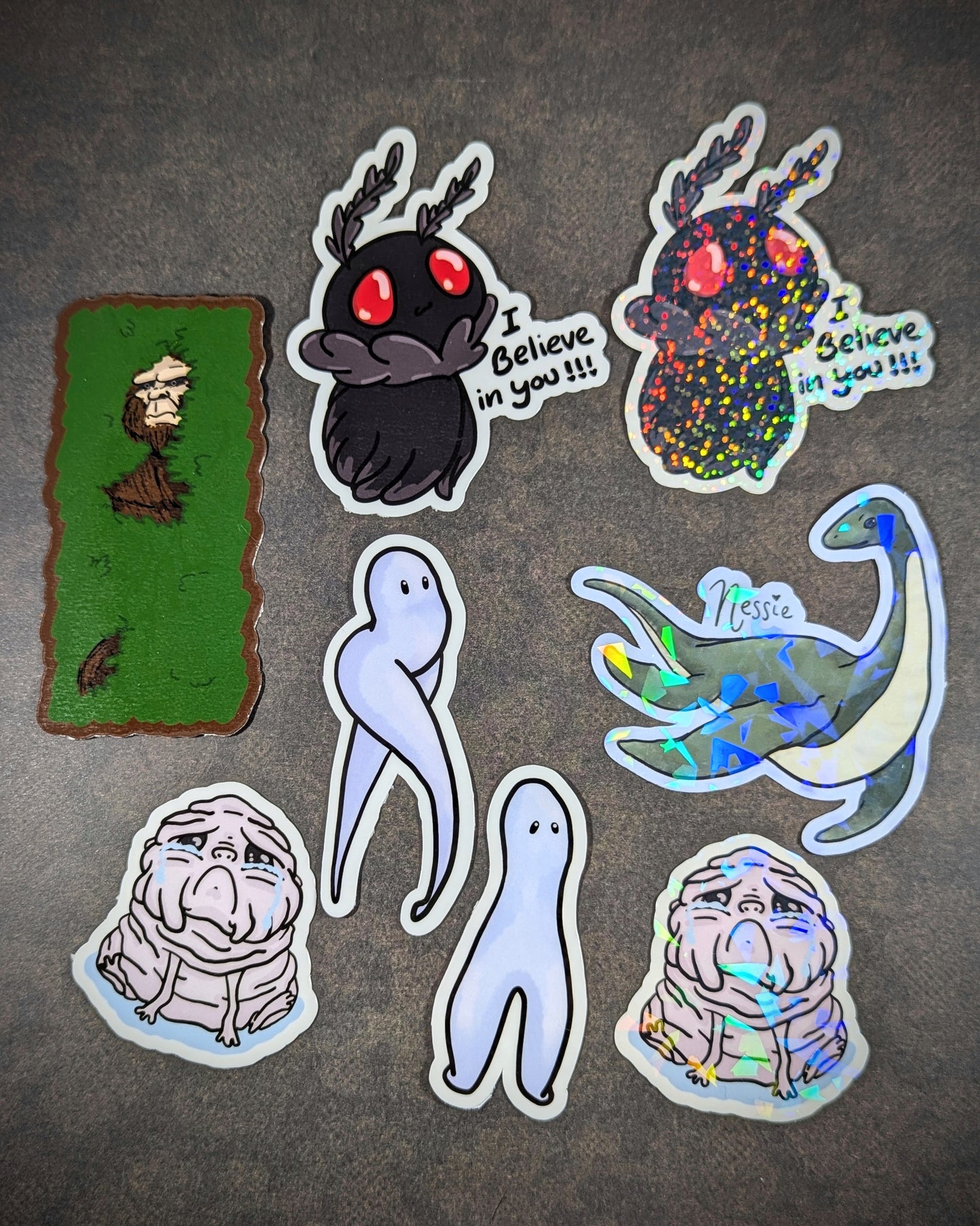 "Assorted cryptid-themed stickers, including Mothman with 'I believe in you' text, holographic Nessie, and other unique, mythical creatures. Perfect for fans of cryptids, folklore, and supernatural decor."