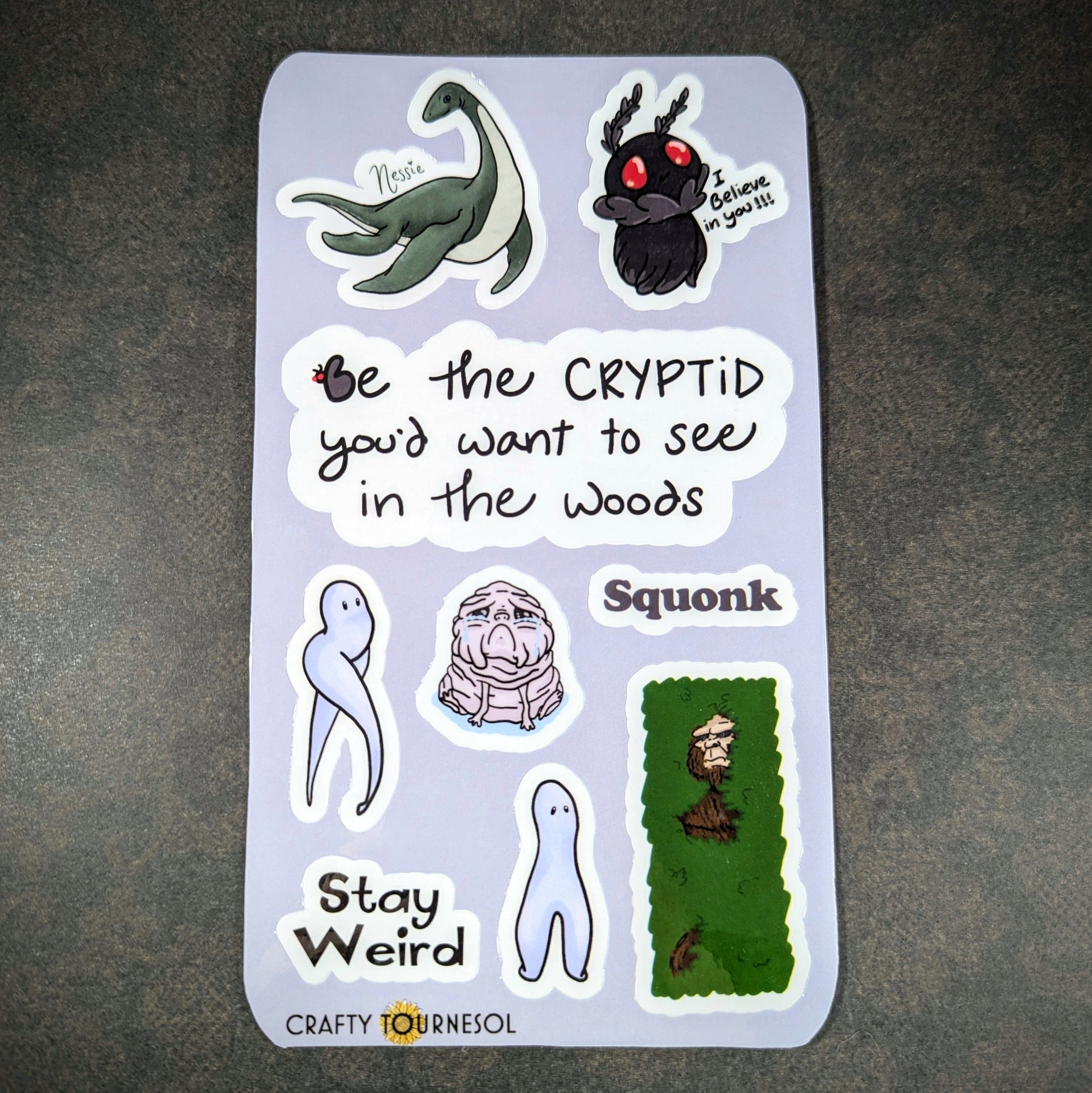 "Cryptid-themed sticker sheet featuring Nessie, Mothman, Squonk, and other quirky designs with phrases like 'Be the cryptid you’d want to see in the woods' and 'Stay Weird' by Crafty Tournesol."