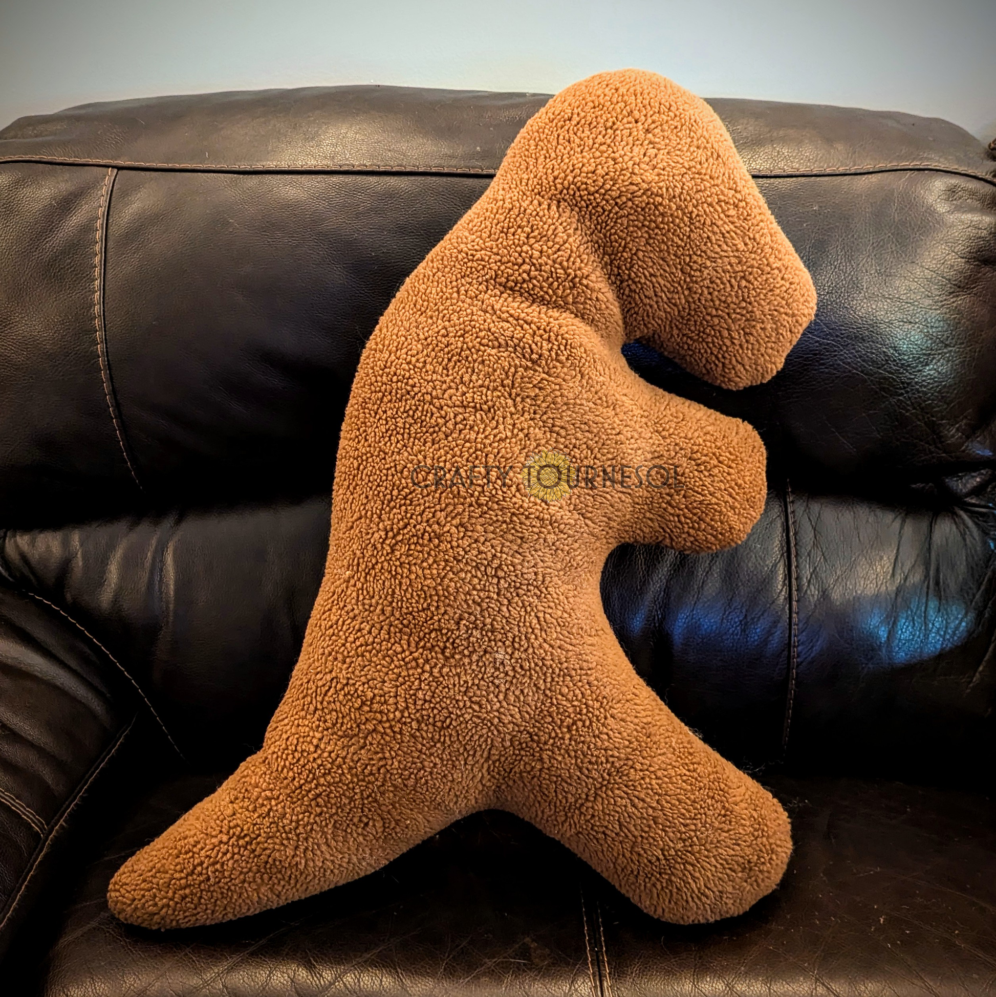 A 3-feet-long, medium brown sherpa pillow in the shape of a t-rex chicken nugget.  Posed on a black leather sofa.