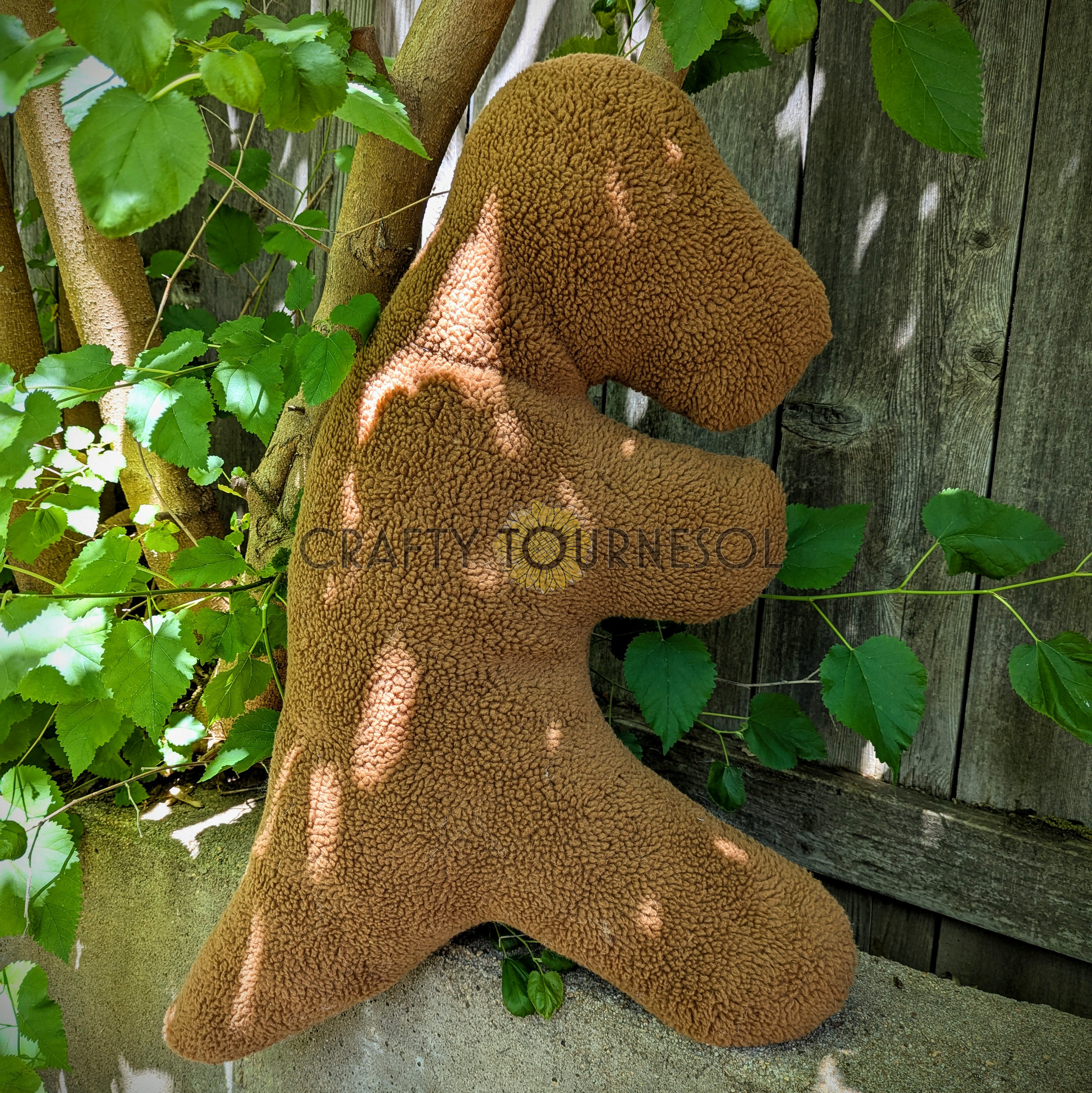 A 3-feet-long, medium brown sherpa pillow in the shape of a t-rex chicken nugget.  Posed in front of a wooden fence and tree.