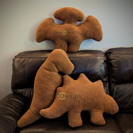 Three 3-feet-long, medium brown sherpa pillows in the shape of dinosaur chicken nuggets.  A t-rex, stegosaurus, and pterodactyl.  Posed on a black leather sofa.