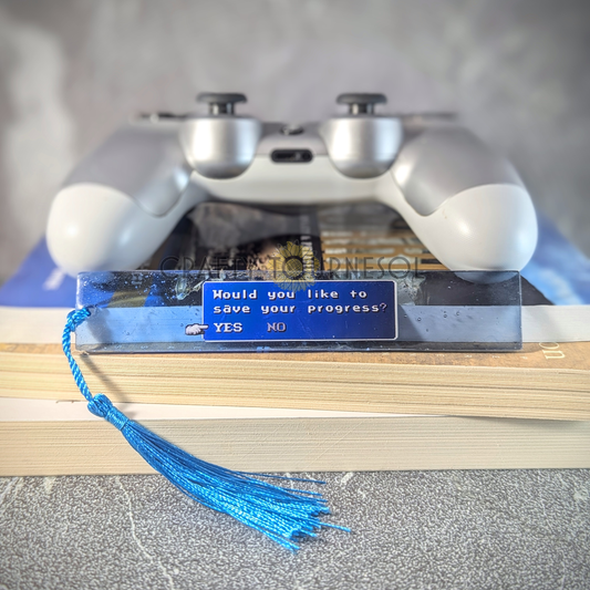 "Pixel art featuring a classic RPG-style save prompt with the text 'Would you like to save your progress? YES NO,' encased in transparent blue resin bookmark, ideal for gamers and nostalgia lovers. Background is a textured gray surface with a book stack and silver Playstation controller."