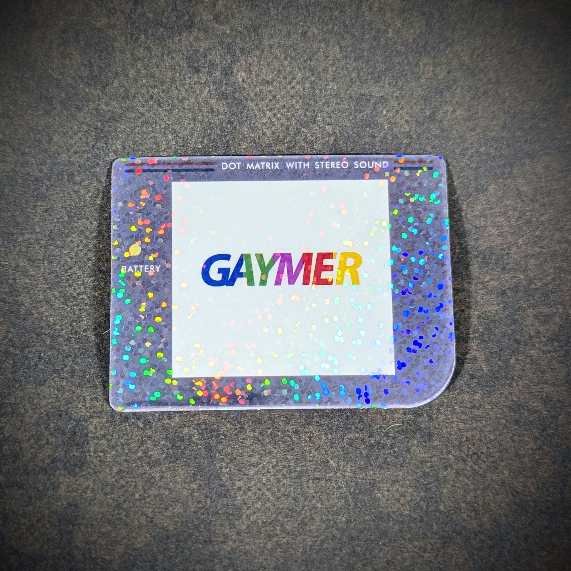 "Holographic sticker with 'Gaymer' in rainbow text on Game Boy screen, inspired by classic Game Boy design with 'Dot Matrix with Stereo Sound' label. A playful, pride-themed decor piece for LGBTQ+ gamers."