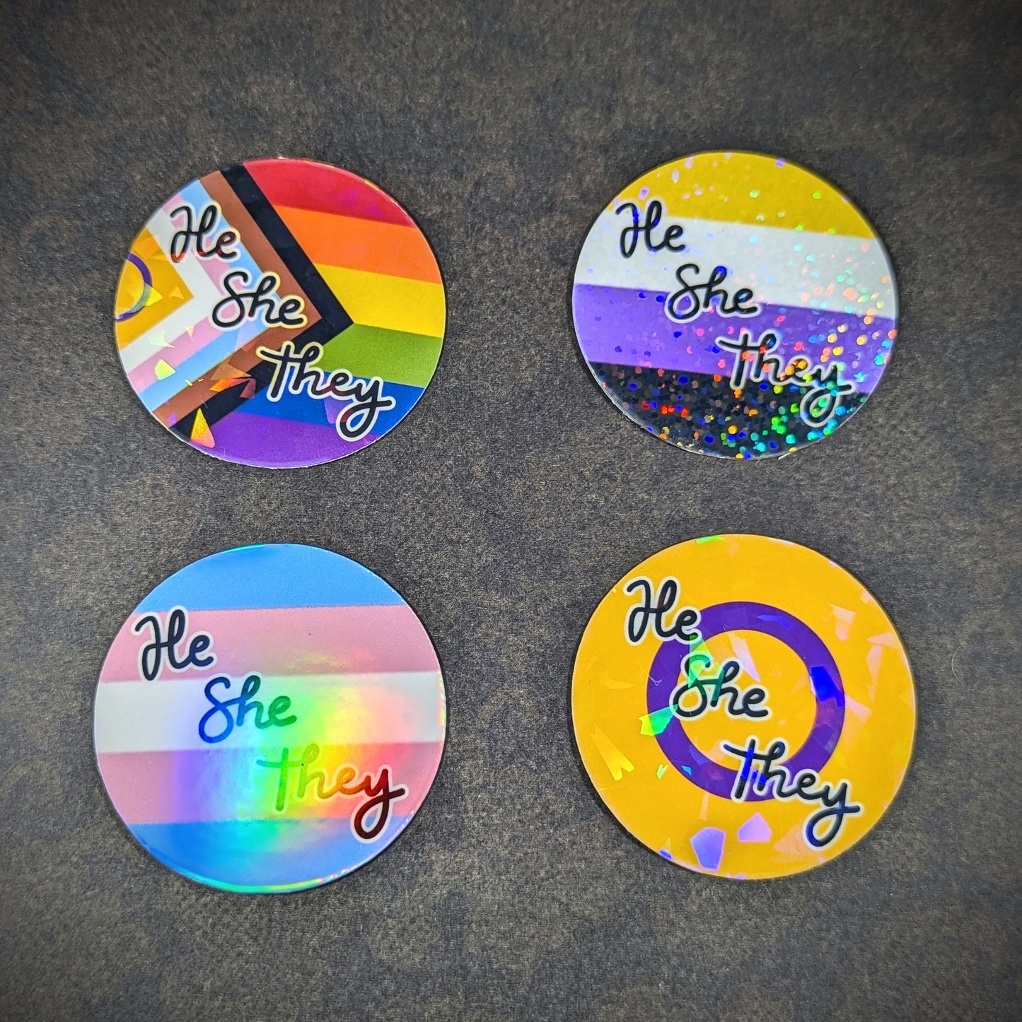 "Round holographic pronoun stickers featuring 'He She They' in various LGBTQ+ pride flag designs, perfect for expressing gender identity and inclusivity on laptops, water bottles, and notebooks."