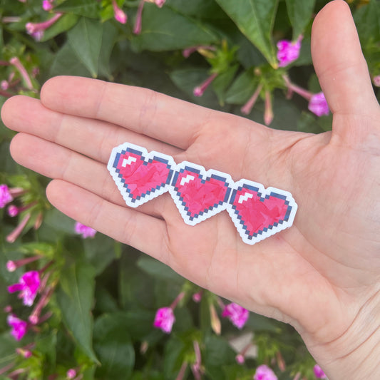 Holographic gamer heart sticker featuring 3 red pixel hearts in a heart container design. Perfect for laptops, water bottles, and game-themed journals.