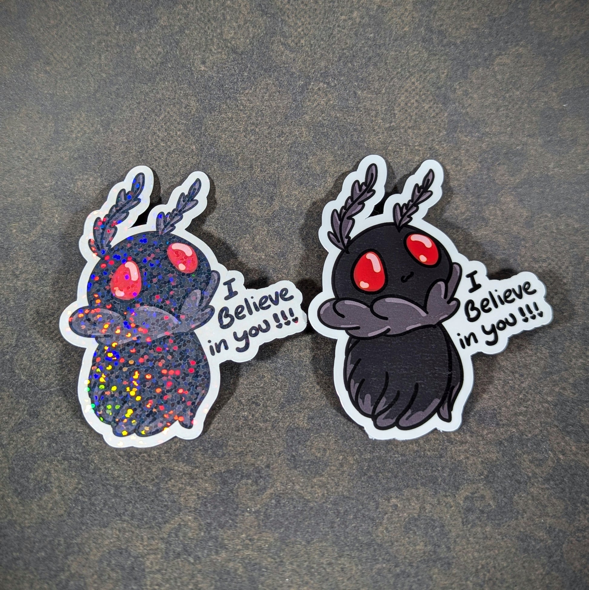 Pair of adorable Mothman stickers with red eyes and 'I Believe in You' text, including a holographic version. Perfect for cryptid fans looking for motivational, cute, and collectible supernatural decor.