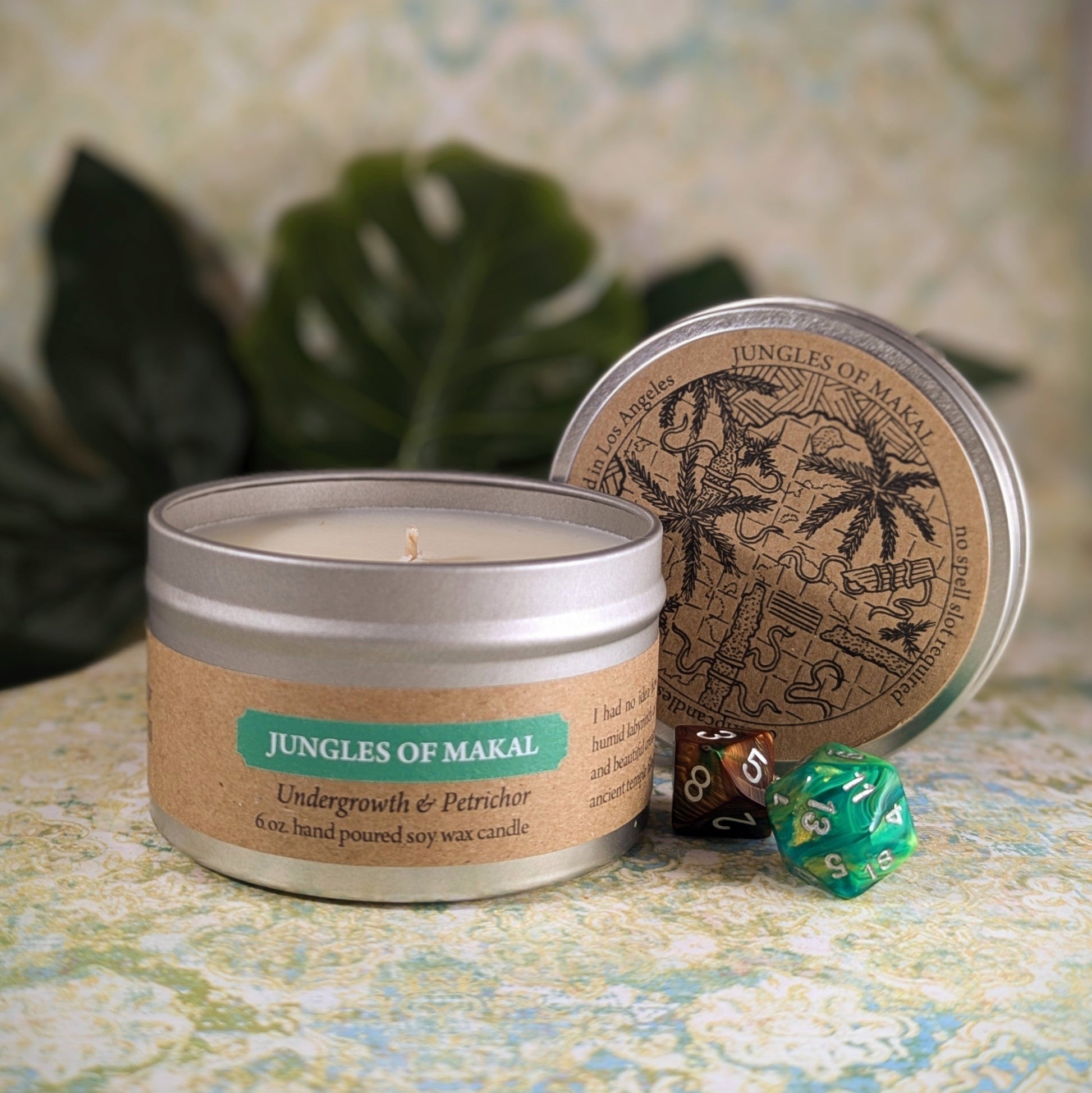 "Jungles of Makal 6 oz soy wax candle tin with Undergrowth & Petrichor scent, staged with RPG dice and tropical decor, perfect for adventurers and fantasy enthusiasts seeking an exotic, jungle-inspired ambiance in their space."