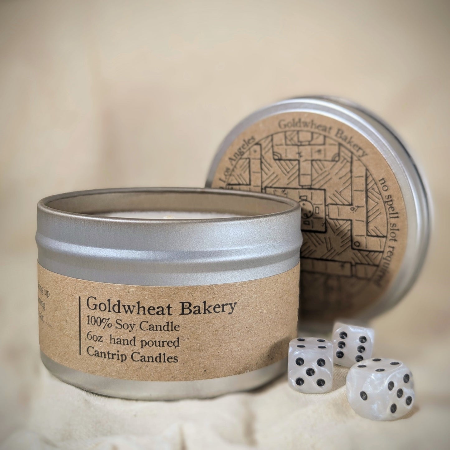 "Goldwheat Bakery 6 oz candle tin, a 100% soy wax blend by Cantrip Candles, with cozy bakery scent, staged with RPG dice and minimalist decor, ideal for gamers and bakery lovers seeking a cozy, bread-scented ambiance for their space."