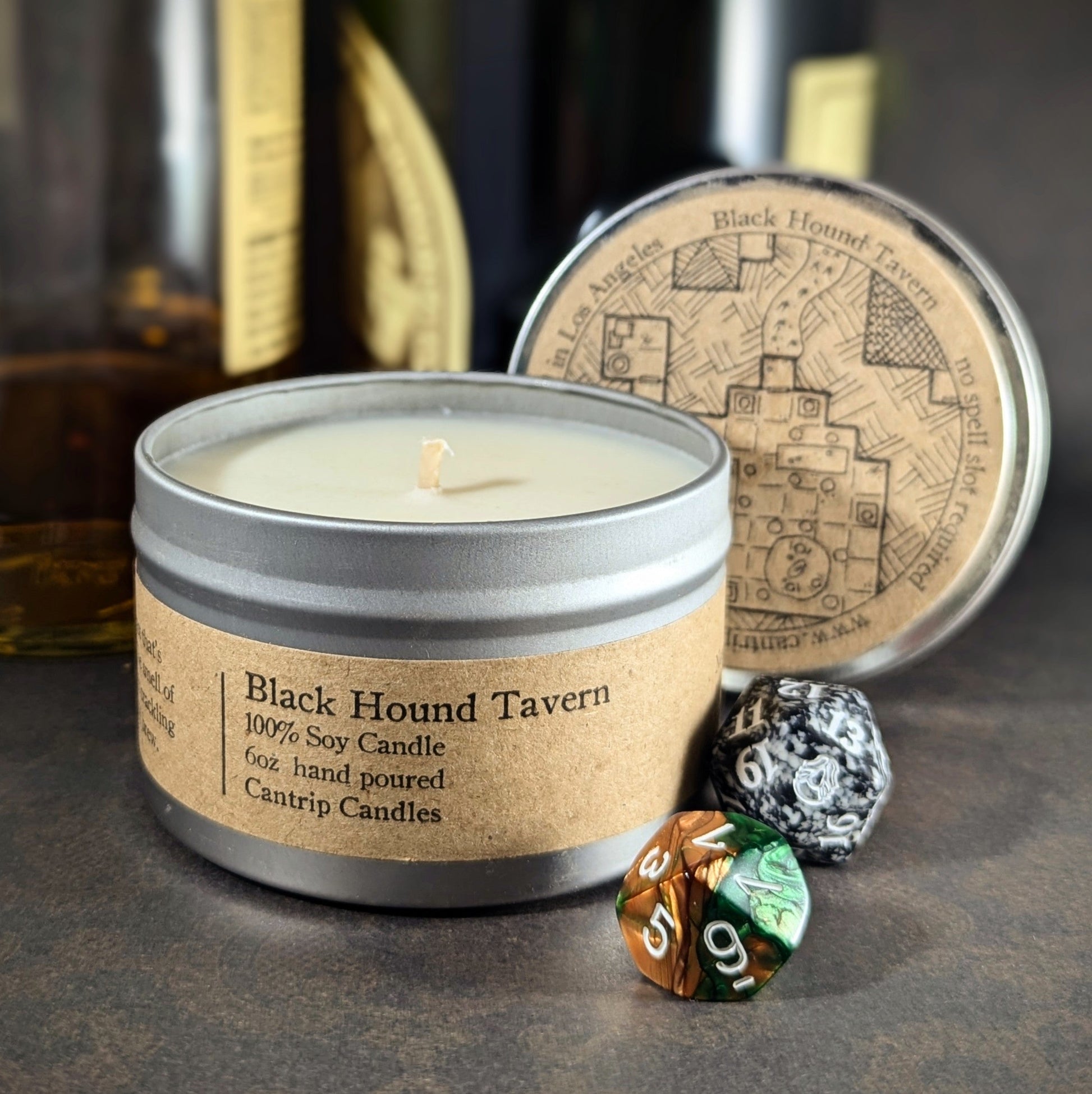 "Black Hound Tavern 6 oz candle tin by Cantrip Candles, a 100% soy wax blend with scents of whiskey and firewood, staged wtih RPG dice and rustic tavern decor, perfect for creating an immersive, cozy atmosphere for gamers and fantasy enthusiasts."