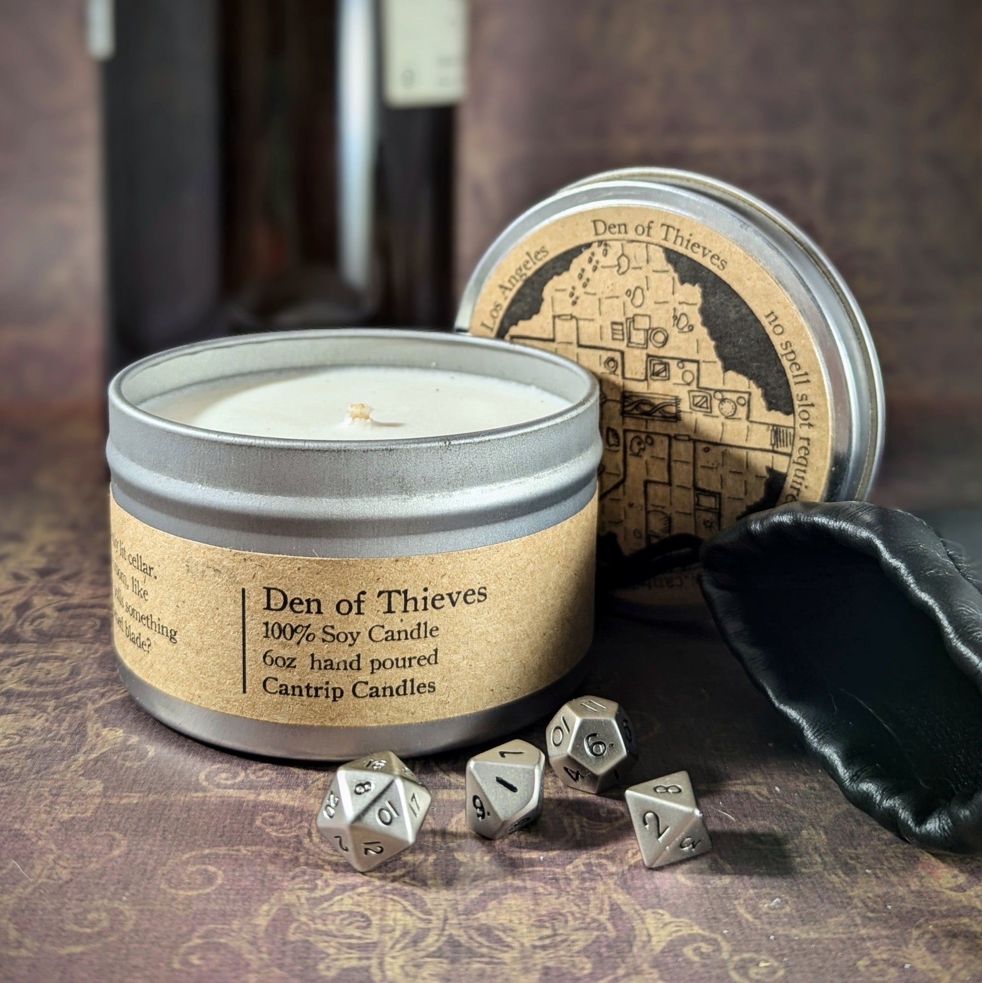 "Den of Thieves 6 oz soy wax candle tin by Cantrip Candles, with smoky notes of red wine, aged leather, and intrigue, staged with RPG dice and dark, mysterious decor, ideal for creating a moody, clandestine atmosphere for gamers and fantasy lovers."