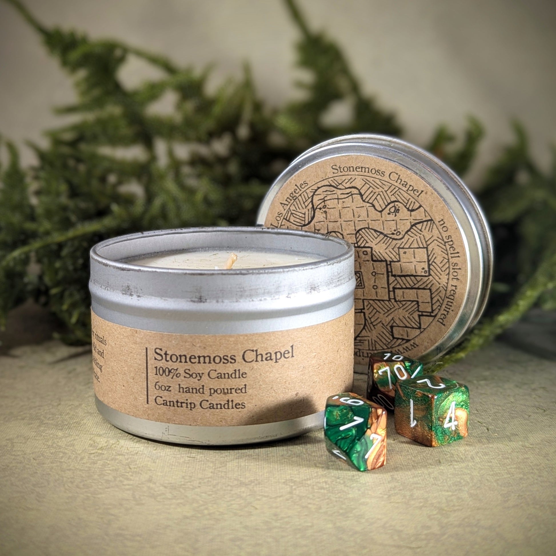 "Stonemoss Chapel 6 oz soy wax candle tin by Cantrip Candles, with scents of stone, moss, and soil, staged with RPG dice and serene, woodland decor, ideal for gamers and fantasy lovers seeking a peaceful, mystical ambiance."