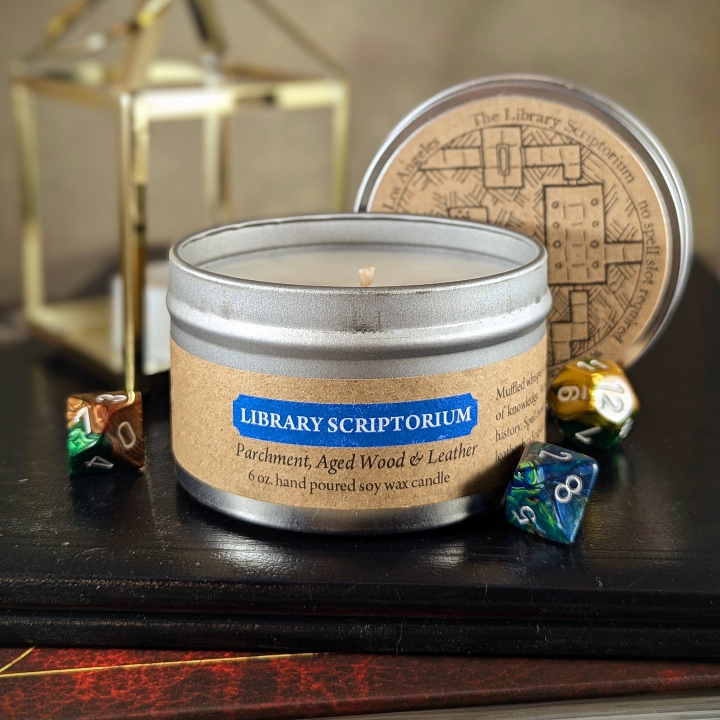 Alt text: "Library Scriptorium 6 oz soy wax candle tin in Parchment, Aged Wood & Leather scent with RPG dice and vintage decor, ideal for book lovers and gamers seeking an old-world library ambiance in their space."