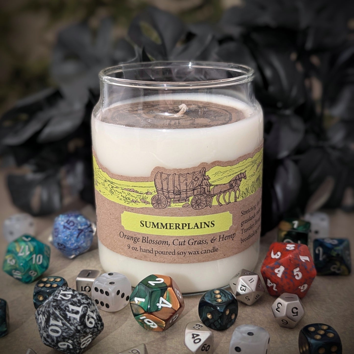 "Summerplains candle by Cantrip Candles, 9 oz soy wax with scents of orange blossom, cut grass, and hemp. Features rustic artwork on label design, staged with RPG dice, evoking open plains and warm summer breezes."