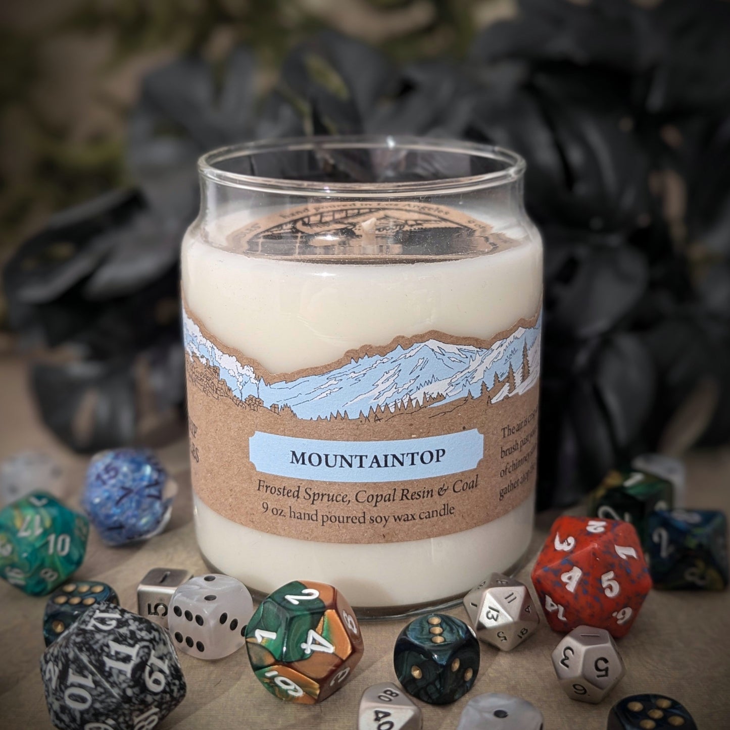 "Mountaintop candle by Cantrip Candles, 9 oz hand-poured soy wax with frosted spruce, copal resin, coal scent. Features scenic mountain illustration on label, staged with RPG dice, perfect for evoking the essence of high altitudes."