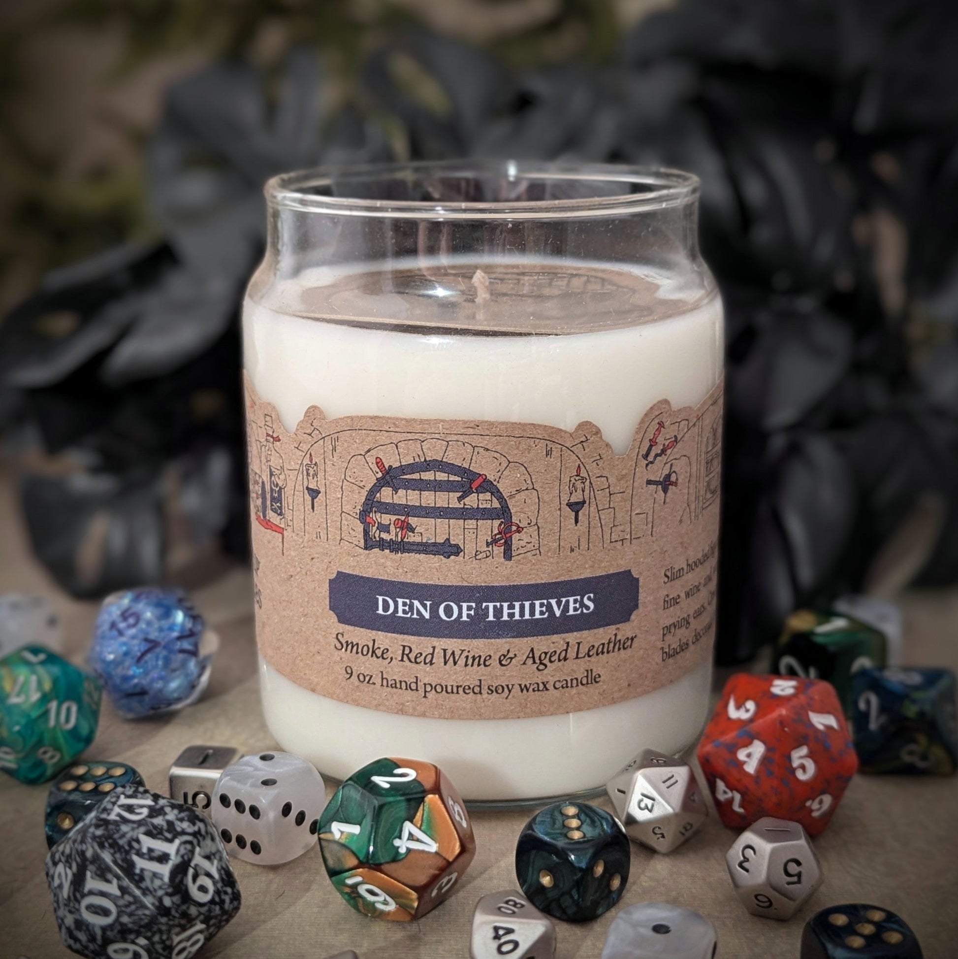 "Den of Thieves candle by Cantrip Candles, 9 oz soy wax with smoky notes of red wine, aged leather, and intrigue, staged with RPG dice. Ideal for ambiance in fantasy tabletop gaming settings."