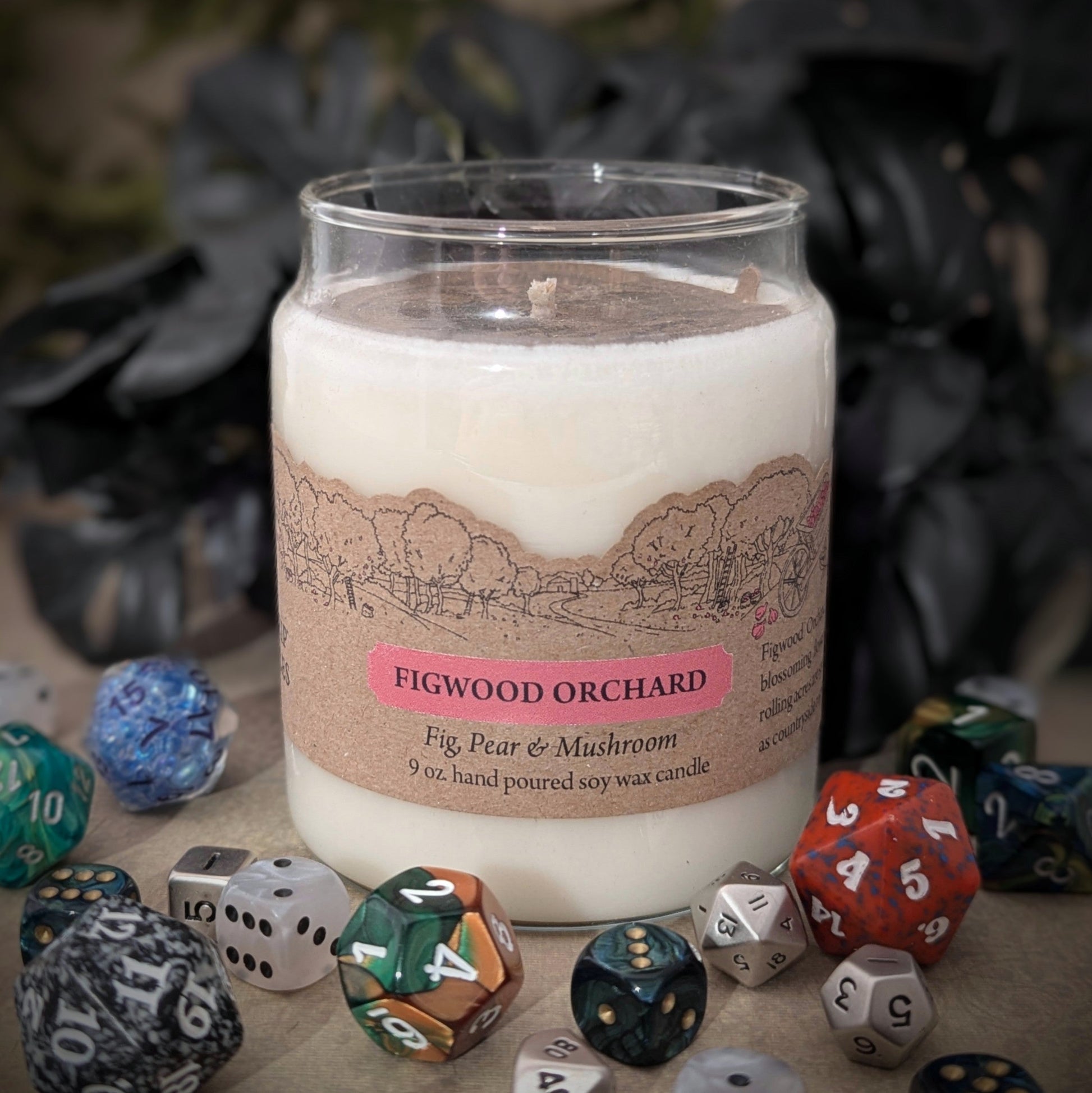 "Figwood Orchard candle by Cantrip Candles, 9 oz soy wax jar, featuring a unique fig, pear, and mushroom scent. Hand-poured with orchard scene label, staged with RPG dice. Perfect for a cozy, nature-inspired ambiance."