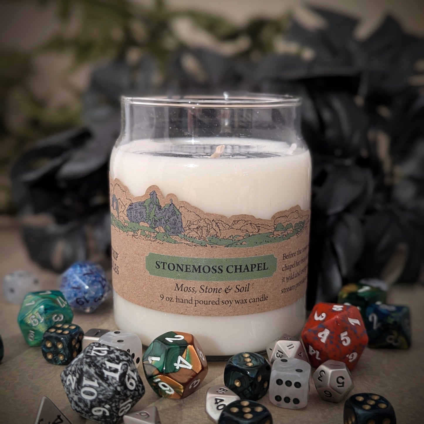 "Stonemoss Chapel candle by Cantrip Candles, 9 oz soy wax with earthy scents of moss, stone, and soil. Features detailed chapel illustration on label, staged with RPG dice, perfect for creating a grounding, nature-inspired ambiance."