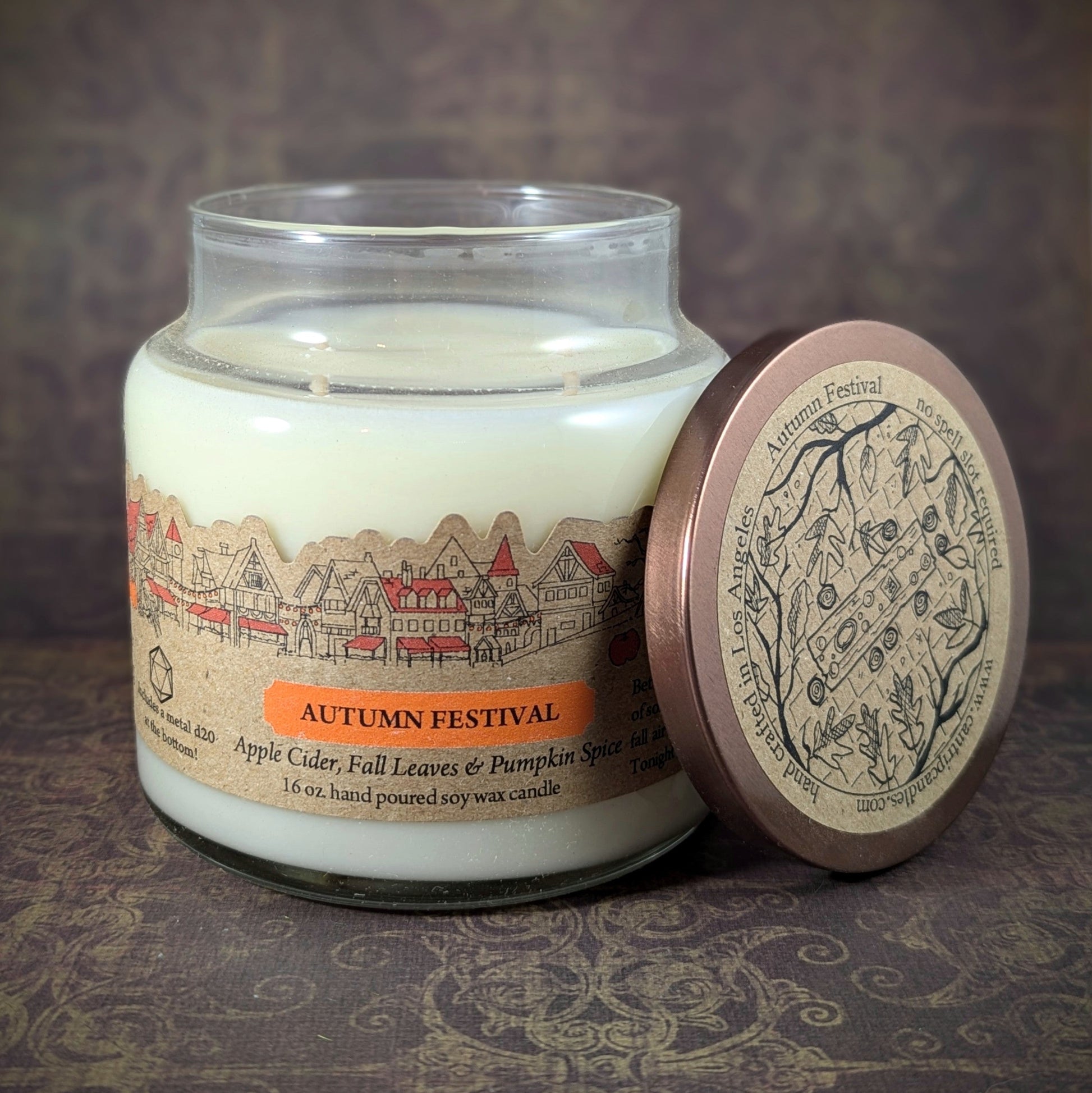 "Autumn Festival candle by Cantrip Candles, 16 oz soy wax, scented with apple cider, fall leaves, and pumpkin spice. Hand-poured with village scene label and intricate lid design, perfect for cozy fall vibes."