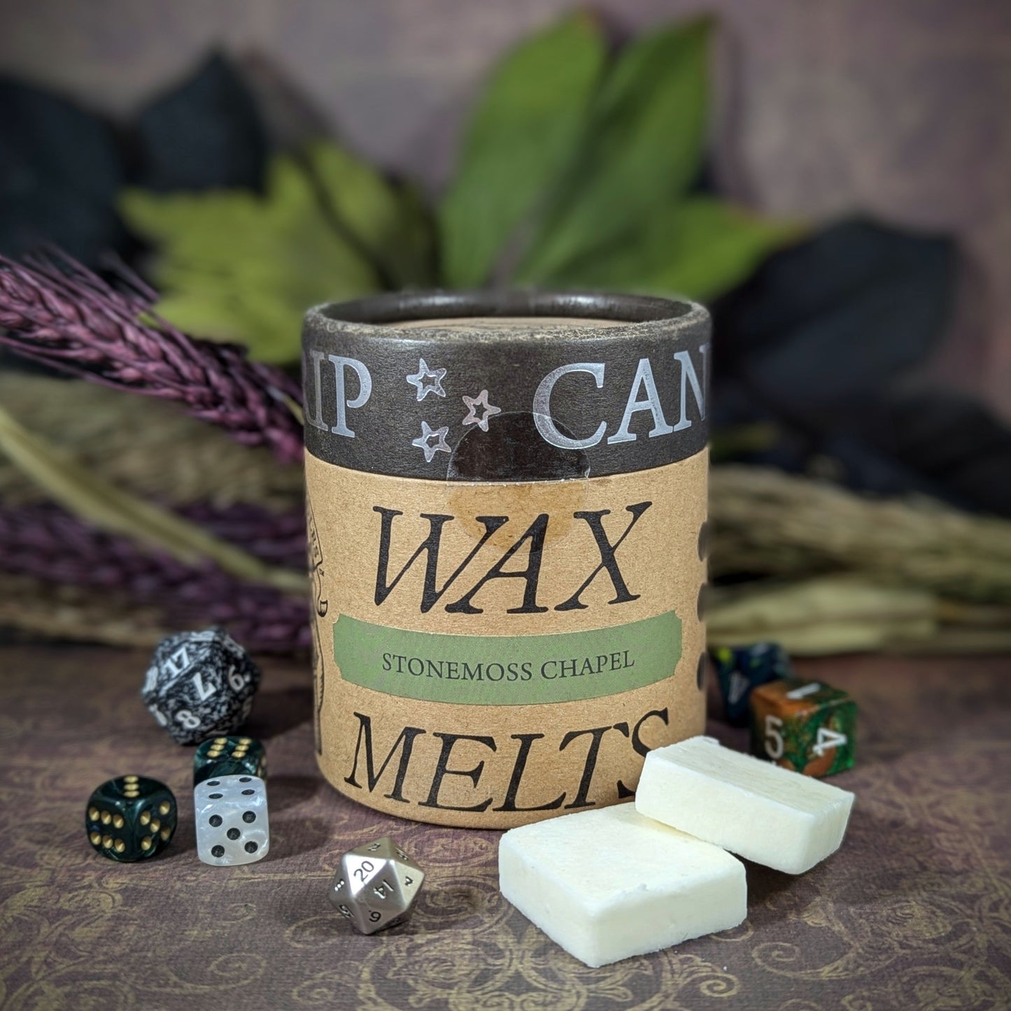 "Cardboard canister of Stonemoss Chapel 5 oz soy wax melts by Cantrip Candles, with earthy scents of moss, stone, and soil, staged with florals and RPG dice. Perfect for creating a grounding, nature-inspired ambiance."