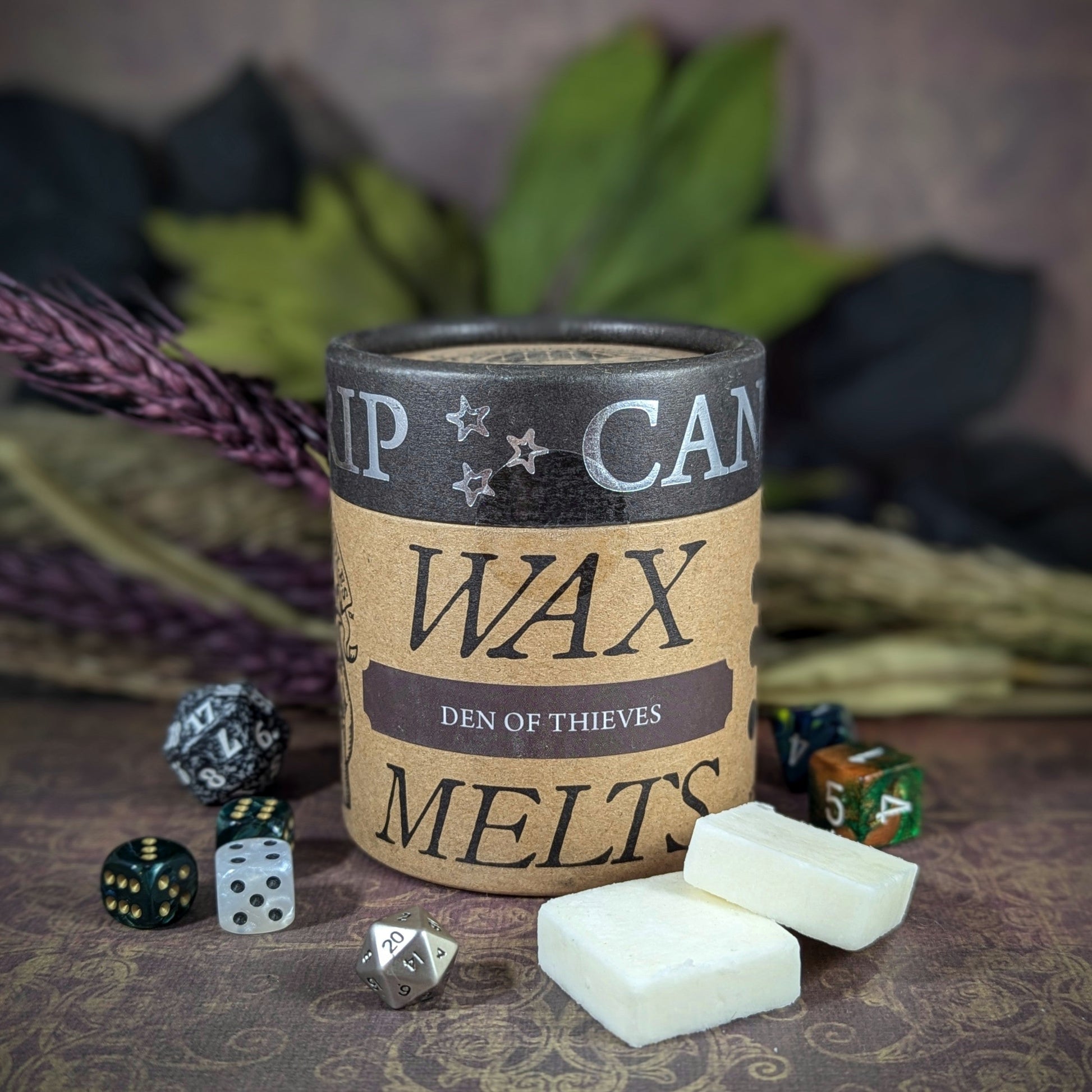 "Cardboard canister of Den of Thieves 5 oz soy wax melts by Cantrip Candles, with smoky notes of red wine, aged leather, and intrigue, staged with florals and RPG dice. Ideal for ambiance in fantasy tabletop gaming settings."