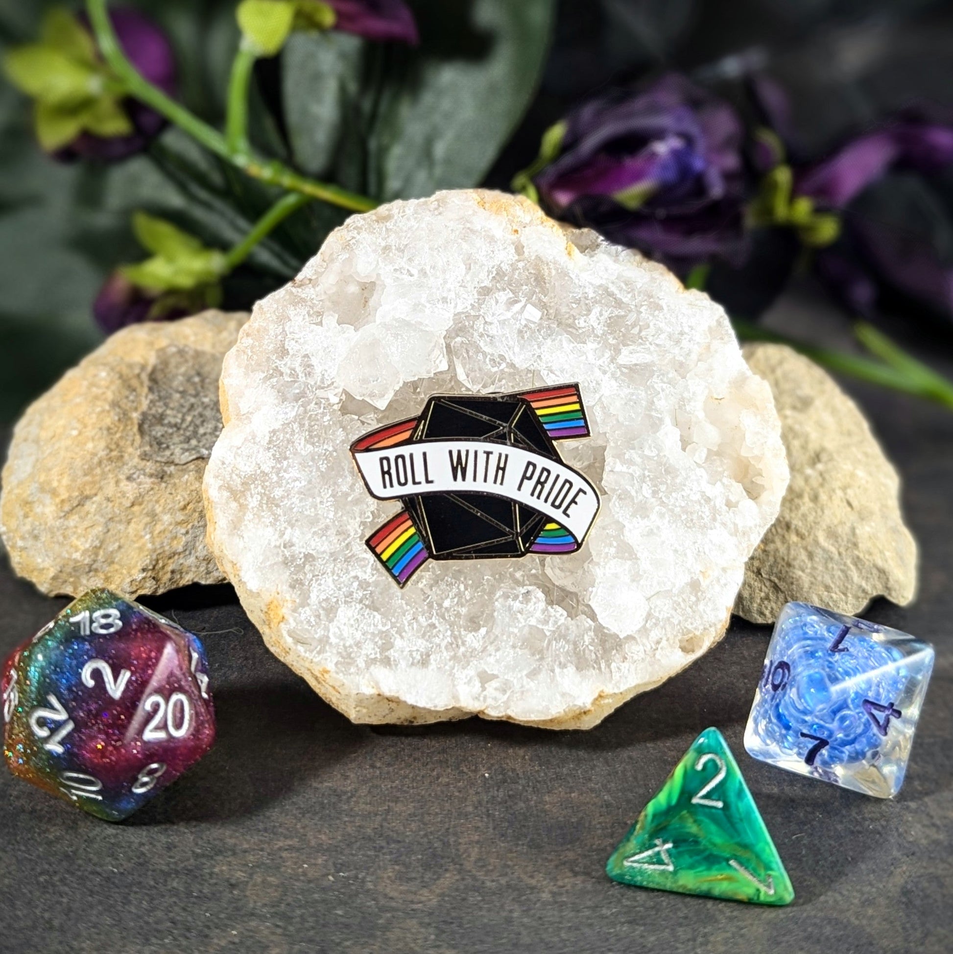 "Enamel pin featuring a black 20-sided die wrapped in a rainbow banner reading, 'Roll With Pride," design by Paola’s Pixels and Cantrip Candles, promoting inclusivity in gaming. Perfect accessory for tabletop RPG fans and LGBTQ+ allies."