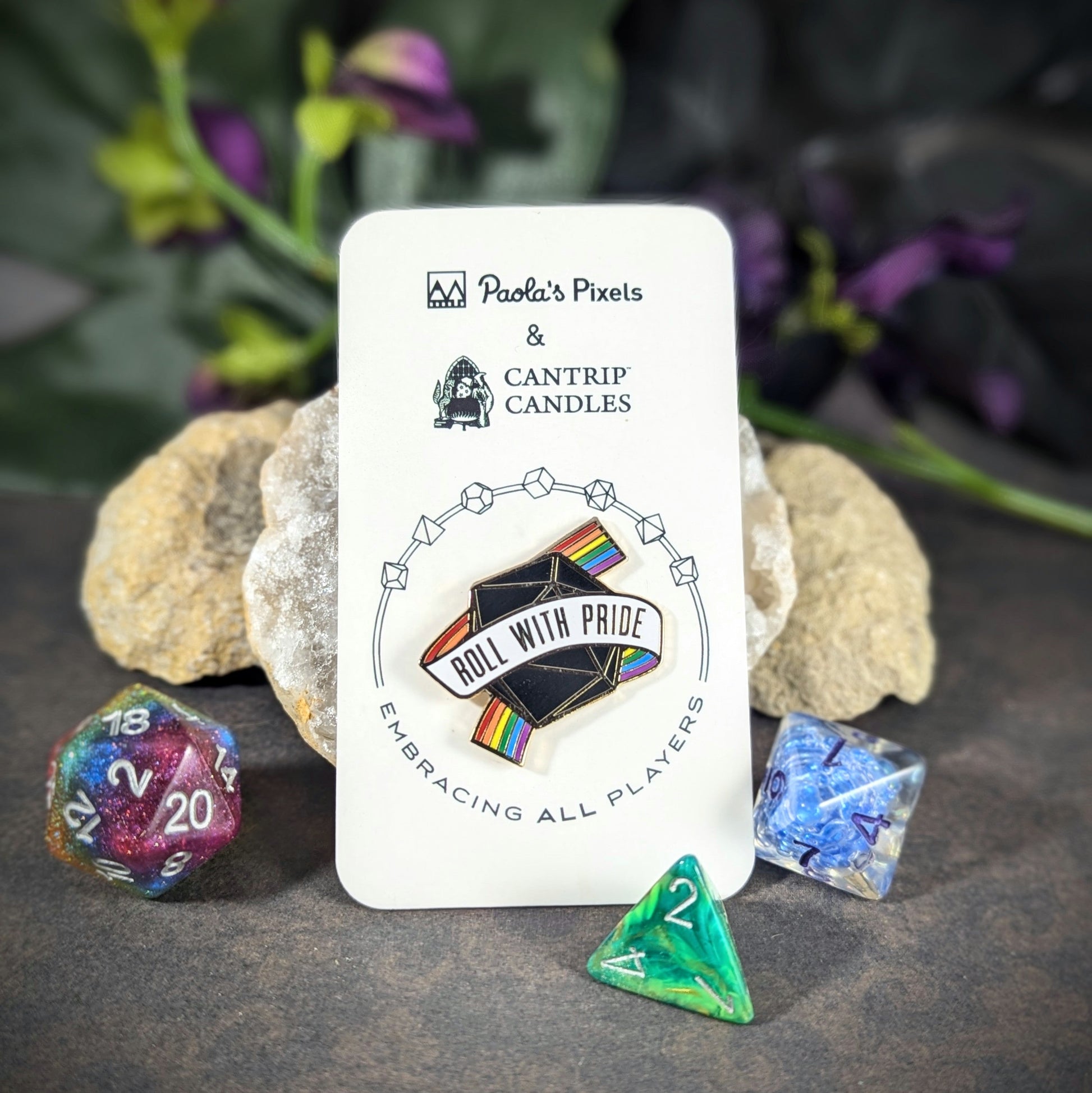 "Enamel pin featuring a black 20-sided die wrapped in a rainbow banner reading, 'Roll With Pride," design by Paola’s Pixels and Cantrip Candles, promoting inclusivity in gaming. Perfect accessory for tabletop RPG fans and LGBTQ+ allies."