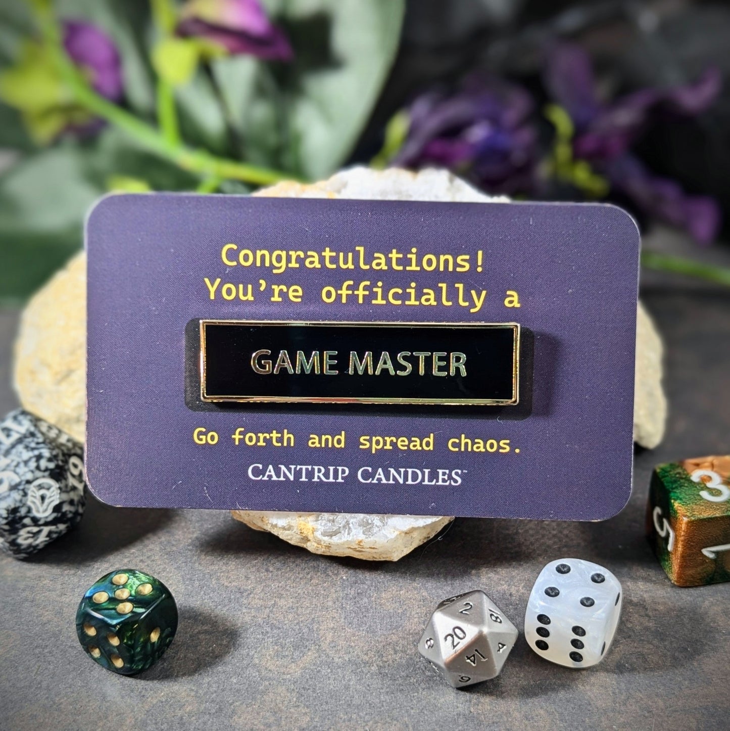 Alt text: "Game Master enamel pin by Cantrip Candles staged with RPG dice, a perfect accessory for tabletop role-playing enthusiasts, celebrating the role of Game Masters with the message: 'Go forth and spread chaos.'"