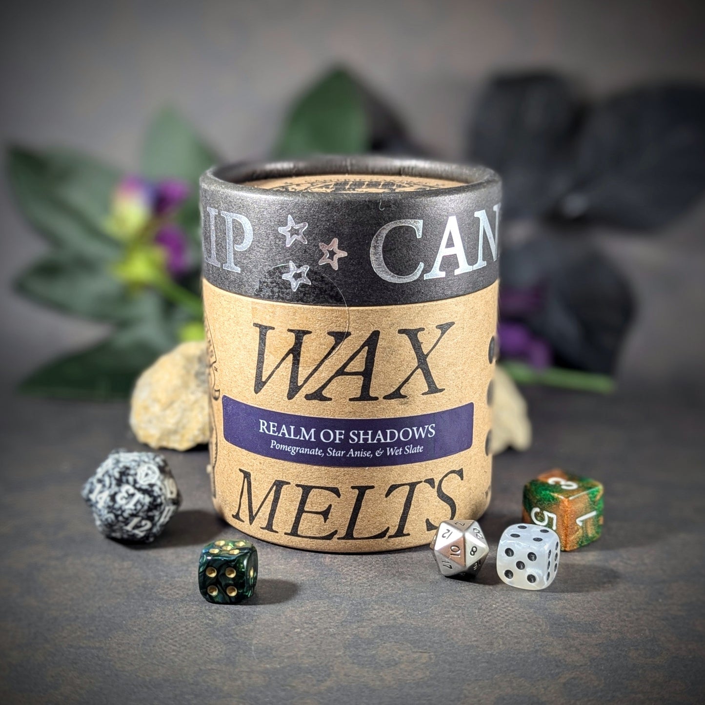 "Cardboard canister of Realm of Shadows 5 oz soy wax melts by Cantrip Candles, with pomegranate, star anise, and wet slate scent, staged with florals and RPG dice. Ideal for those seeking a mysterious, immersive fragrance."