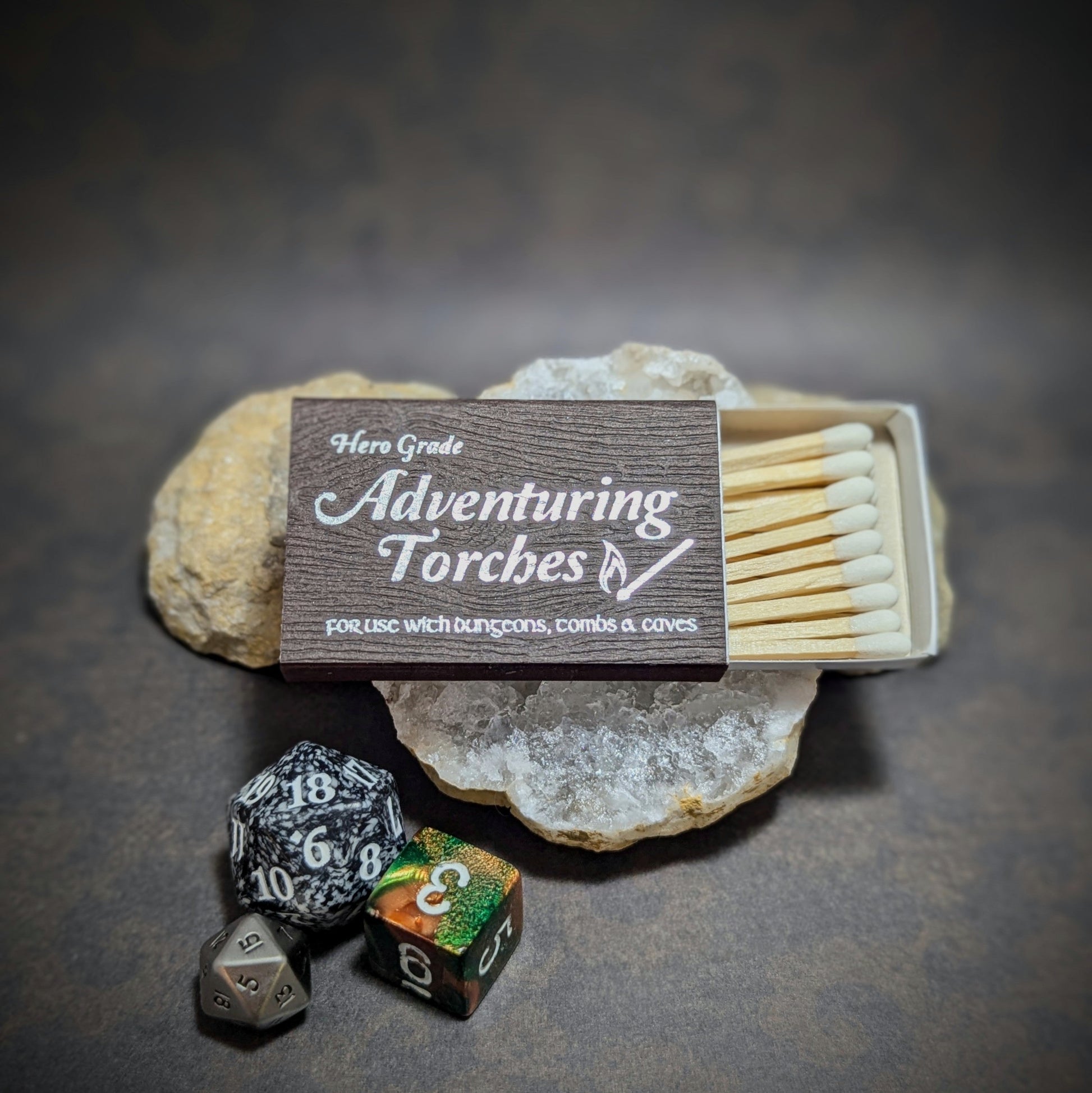 "Hero Grade Adventuring Torches matchbox by Cantrip Candles, designed for use in dungeons, tombs, and caves, with RPG dice and stone decor, ideal for fantasy gamers and tabletop role-playing sessions."