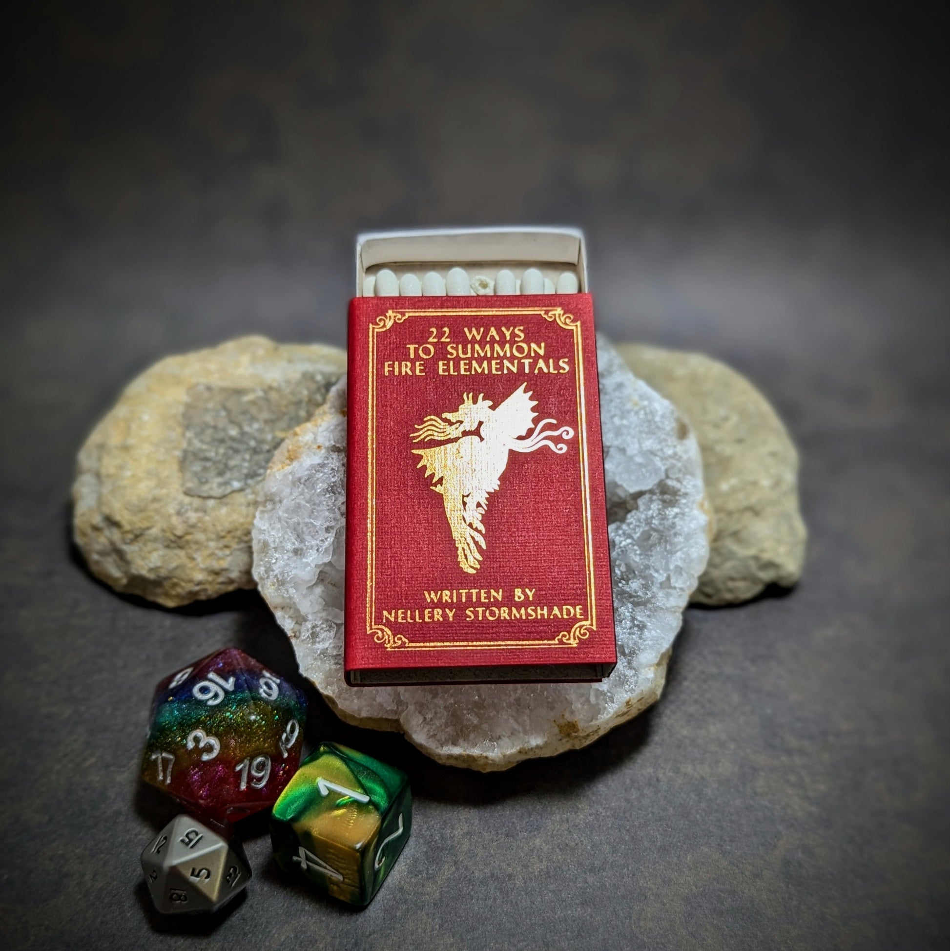 "22 Ways to Summon Fire Elementals matchbox by Cantrip Candles, styled as a magical book with red and gold design, accompanied by RPG dice, perfect for fantasy enthusiasts and tabletop gamers."