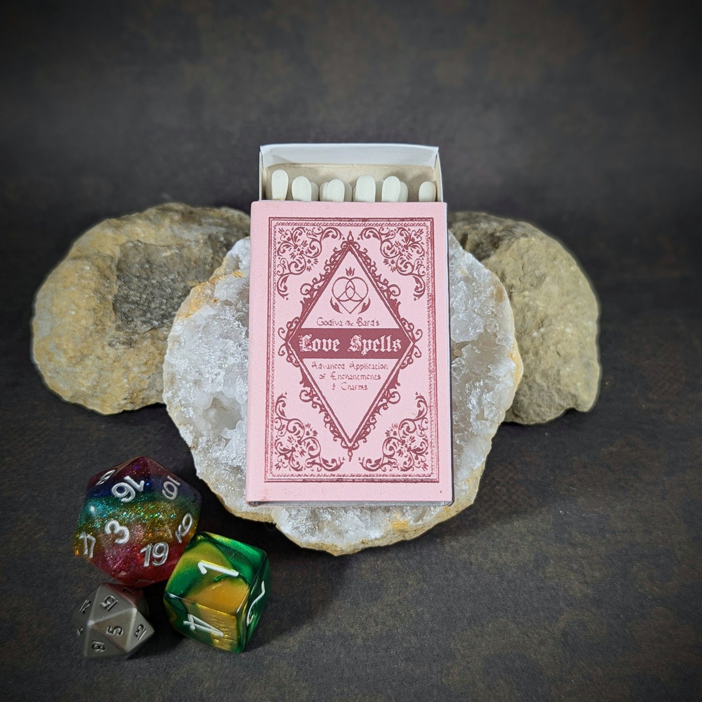 "Love Spells matchbox by Cantrip Candles, styled as a vintage pink spellbook with intricate design, accompanied by RPG dice, perfect for fantasy enthusiasts and tabletop gamers seeking a romantic, mystical touch."