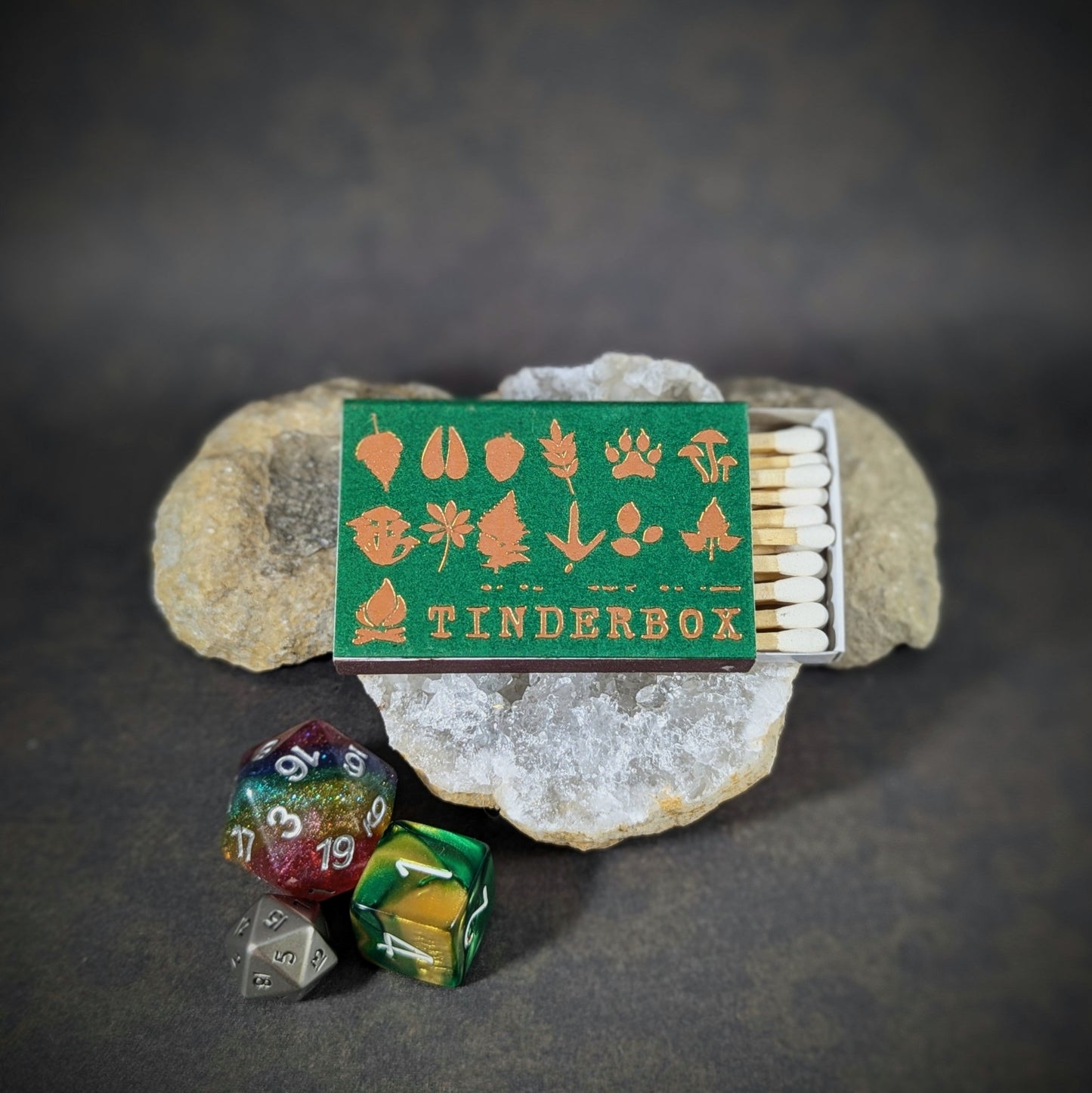 "Tinderbox matchbox by Cantrip Candles, featuring nature-inspired icons in orange on a green background, accompanied by RPG dice, perfect for adventurers and fantasy enthusiasts seeking a rustic accessory."