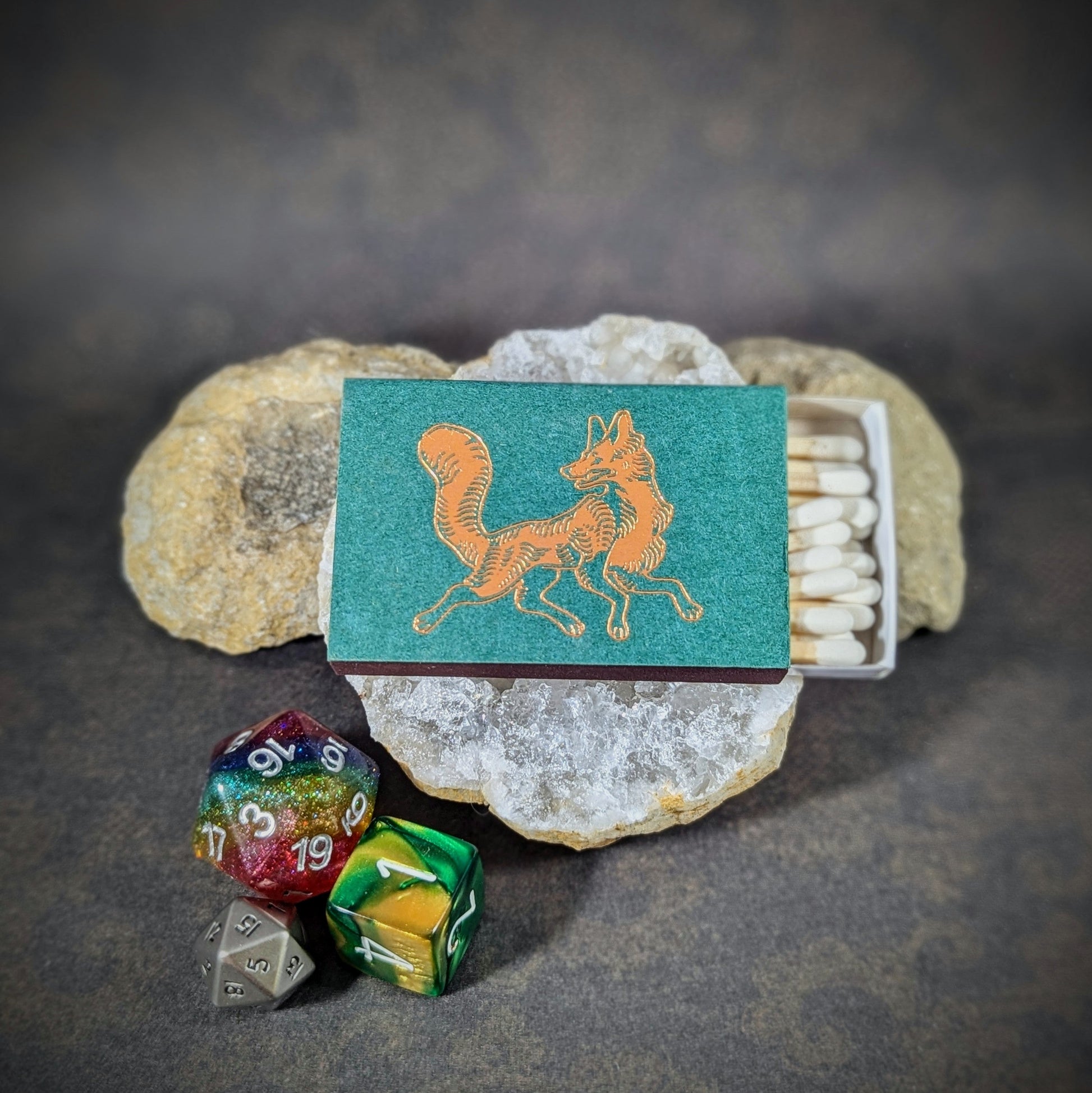 "Matchbox by Cantrip Candles featuring an orange fox design on a green background, accompanied by RPG dice, perfect for fantasy enthusiasts and gamers looking for a unique, woodland-themed accessory."