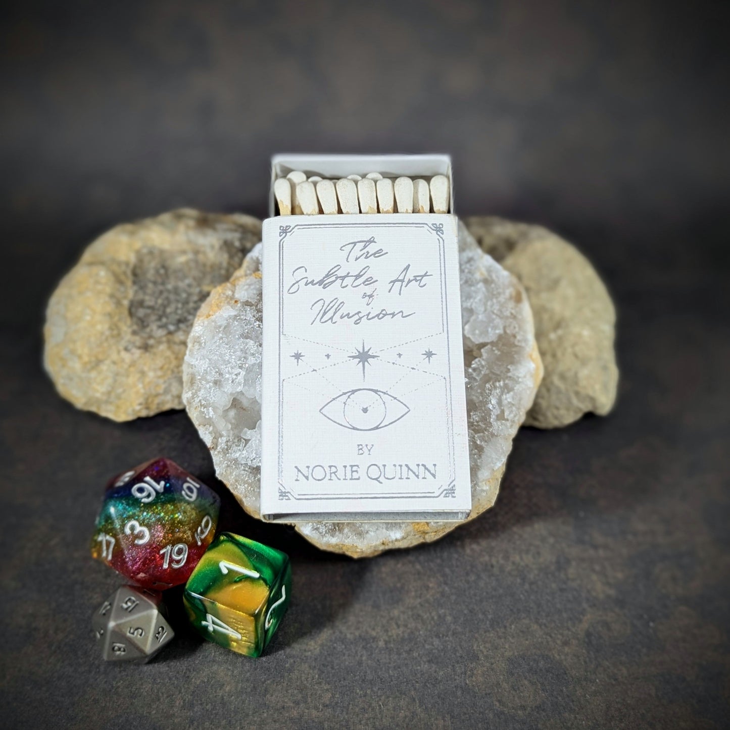 "The Subtle Art of Illusion matchbox by Cantrip Candles, designed as a mystical book cover with an eye symbol, accompanied by RPG dice, ideal for fantasy enthusiasts and tabletop gamers."