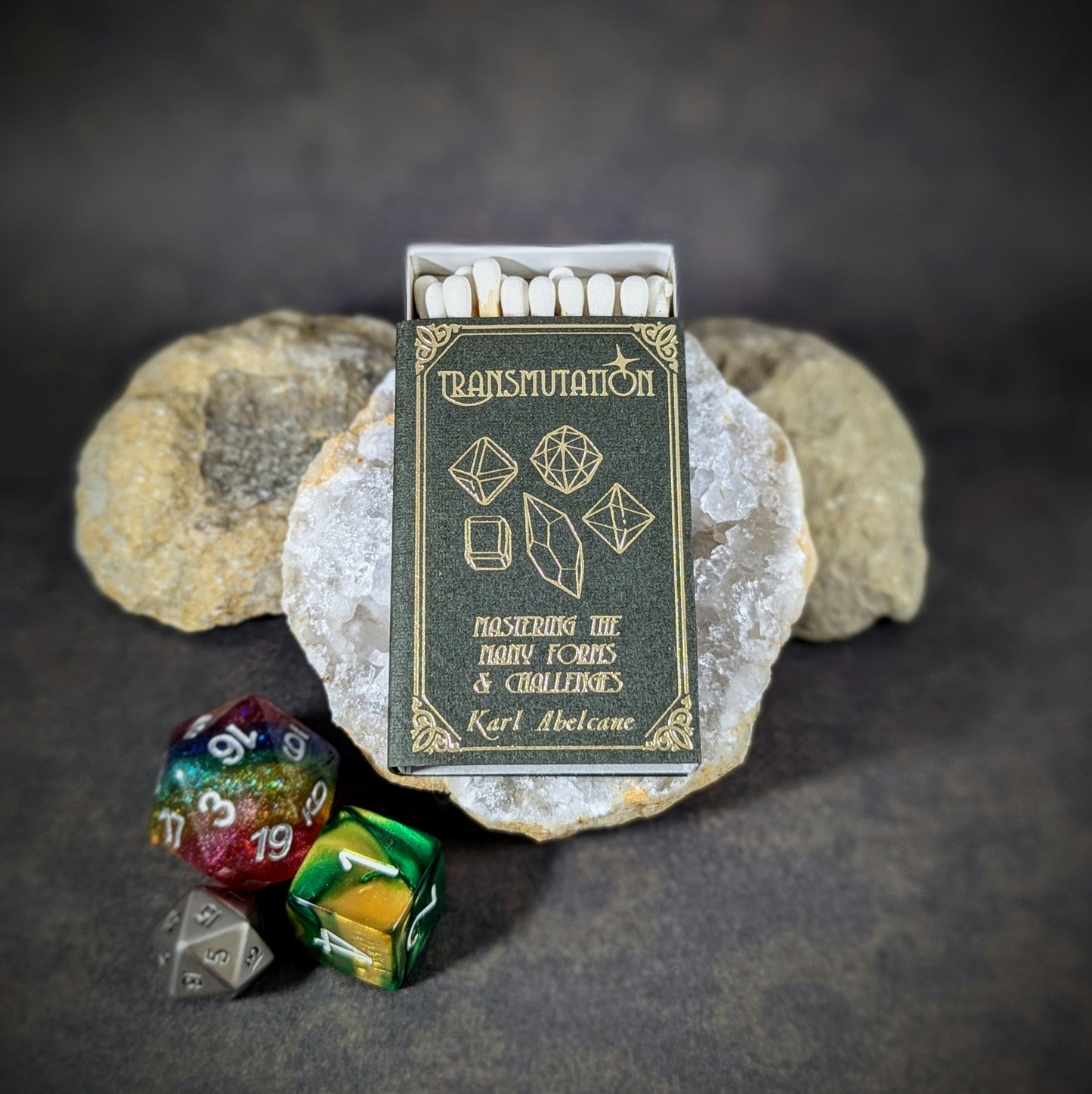 "Transmutation matchbox by Cantrip Candles, designed as a spellbook with gold geometric shapes on a dark background, accompanied by RPG dice, ideal for fantasy enthusiasts and tabletop gamers."