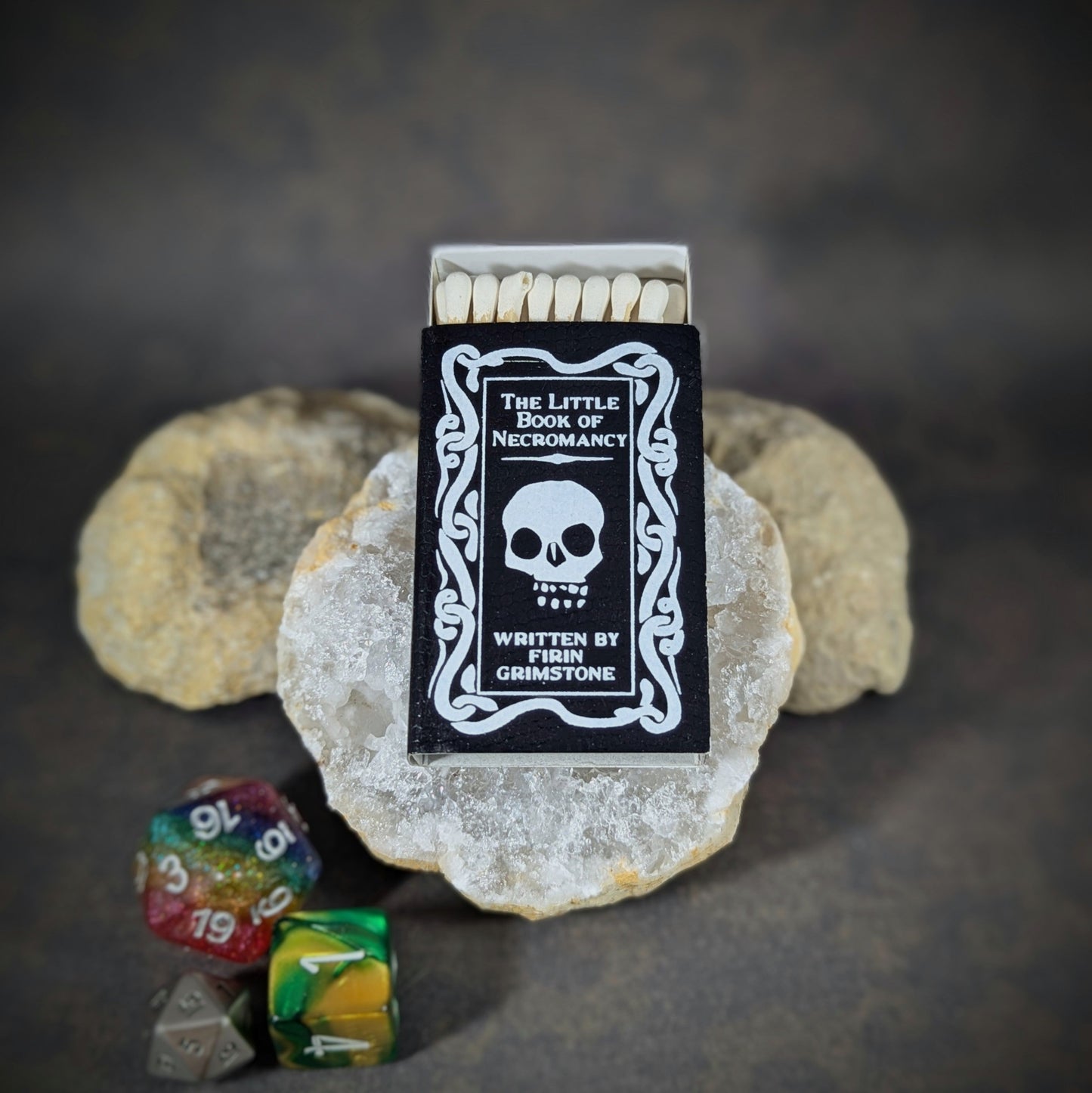 "The Little Book of Necromancy matchbox by Cantrip Candles, styled as a black spellbook with a skull design, accompanied by RPG dice, perfect for fantasy enthusiasts and dark-themed tabletop gaming."