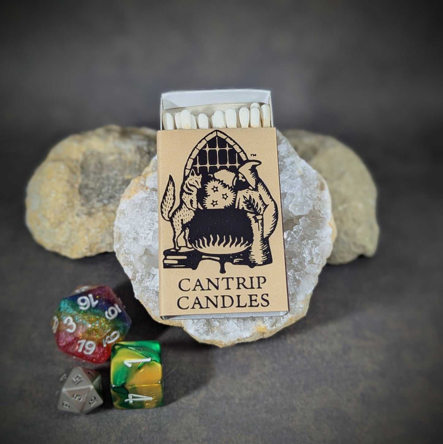 "Cantrip Candles matchbox with whimsical artwork and RPG dice, set against a stone background, perfect for fantasy gamers and candle enthusiasts seeking a magical, role-playing ambiance."