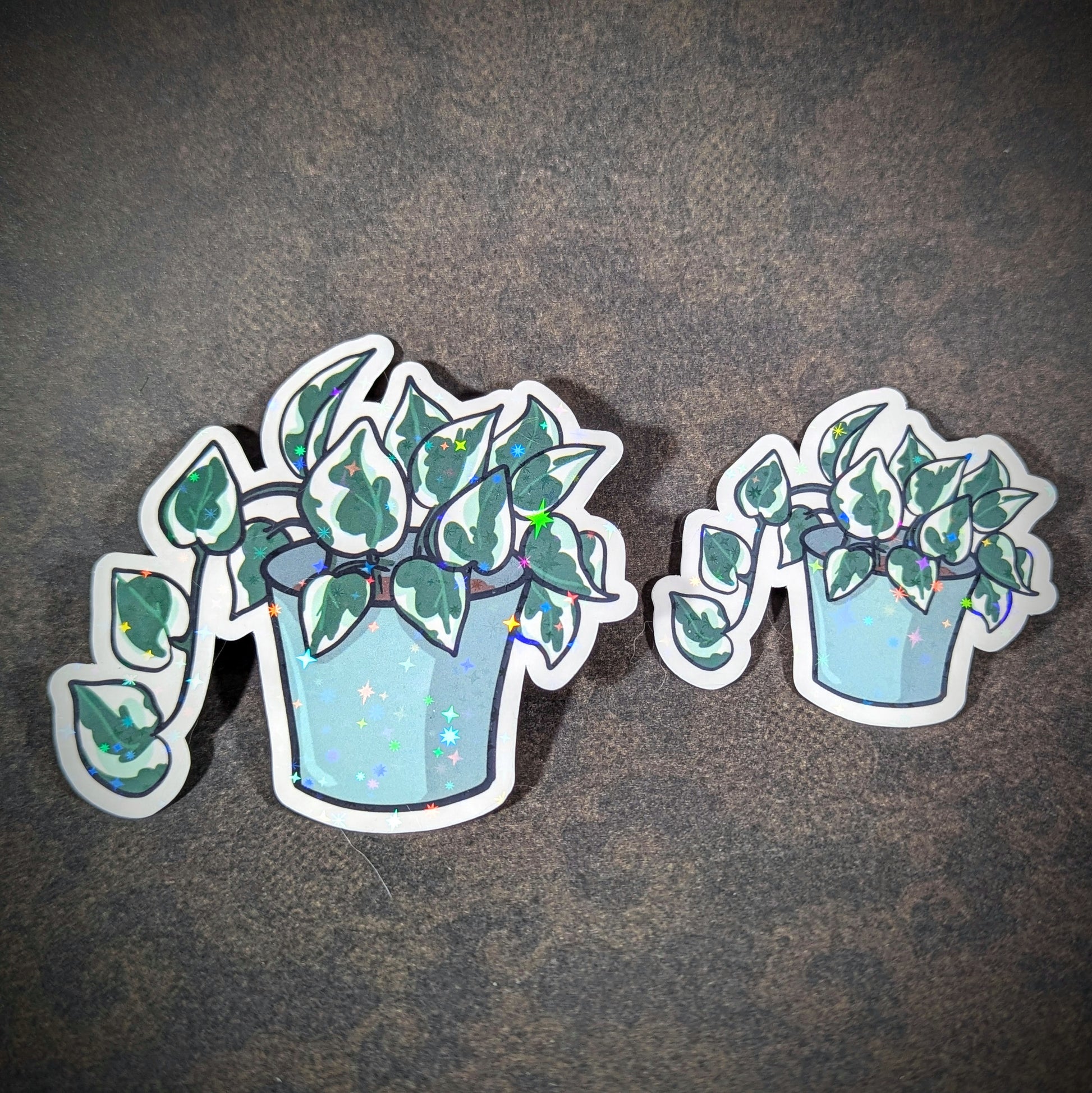 "Pair of holographic pothos N'Joy plant stickers in blue pots, featuring trailing green and white leaves with sparkling accents. Ideal for plant lovers adding a touch of greenery to laptops, journals, or decor."