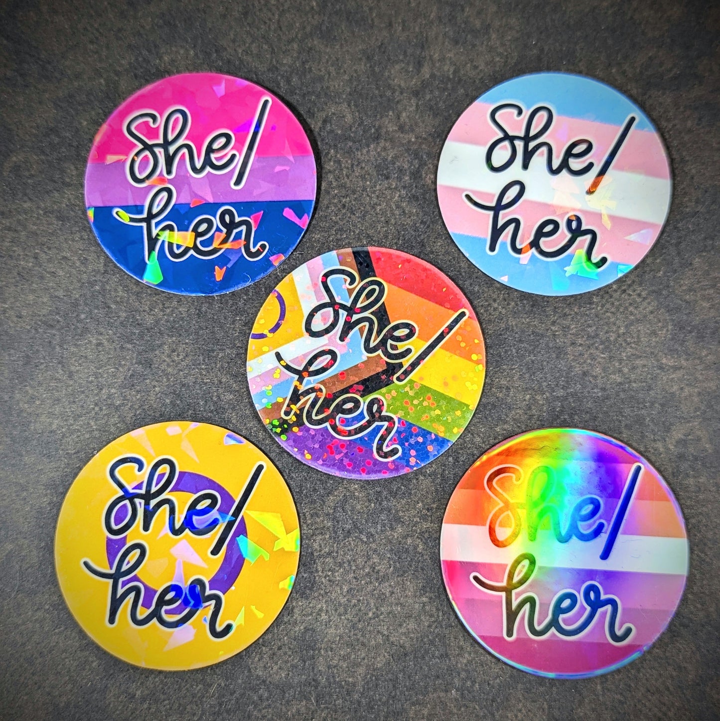 "Round holographic pronoun stickers featuring 'She her' in various LGBTQ+ pride flag designs, perfect for expressing gender identity and inclusivity on laptops, water bottles, and notebooks."